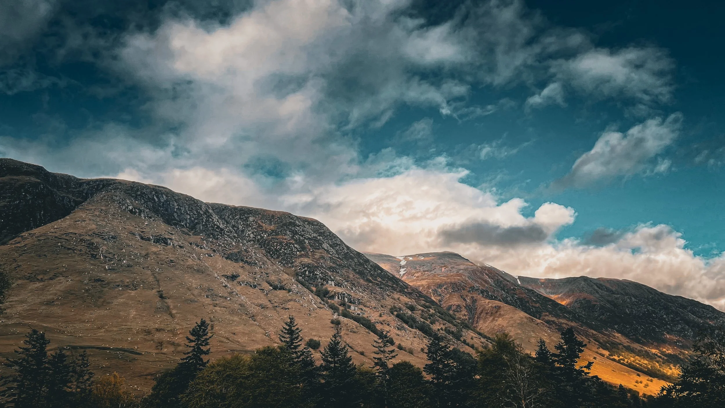 Mountain landscape with brown rocky slopes and sparse vegetation, under a partly cloudy sky.
