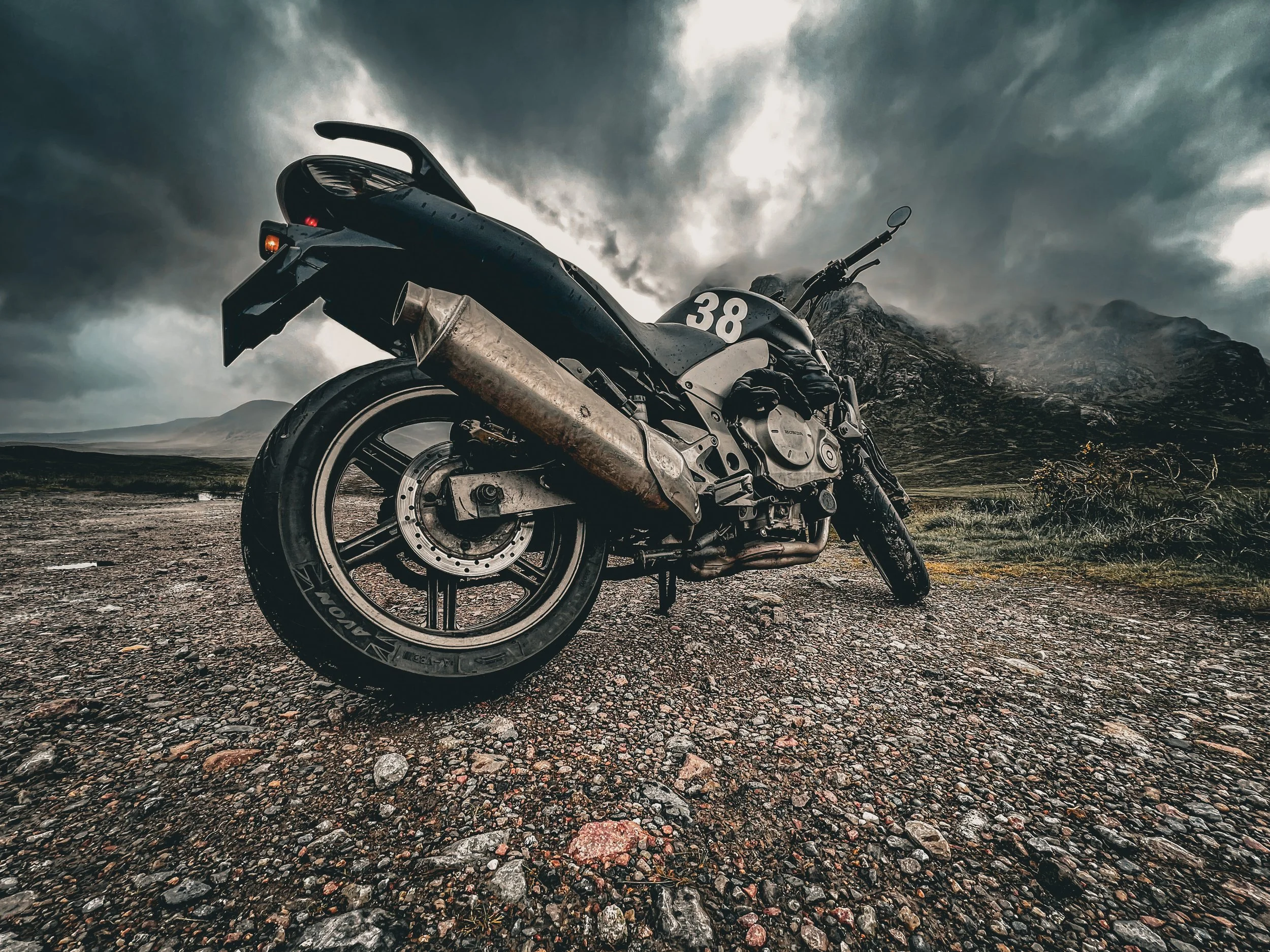 A black motorcycle with a number 38 on the side parked on a gravel surface with mountains and a cloudy sky in the background.