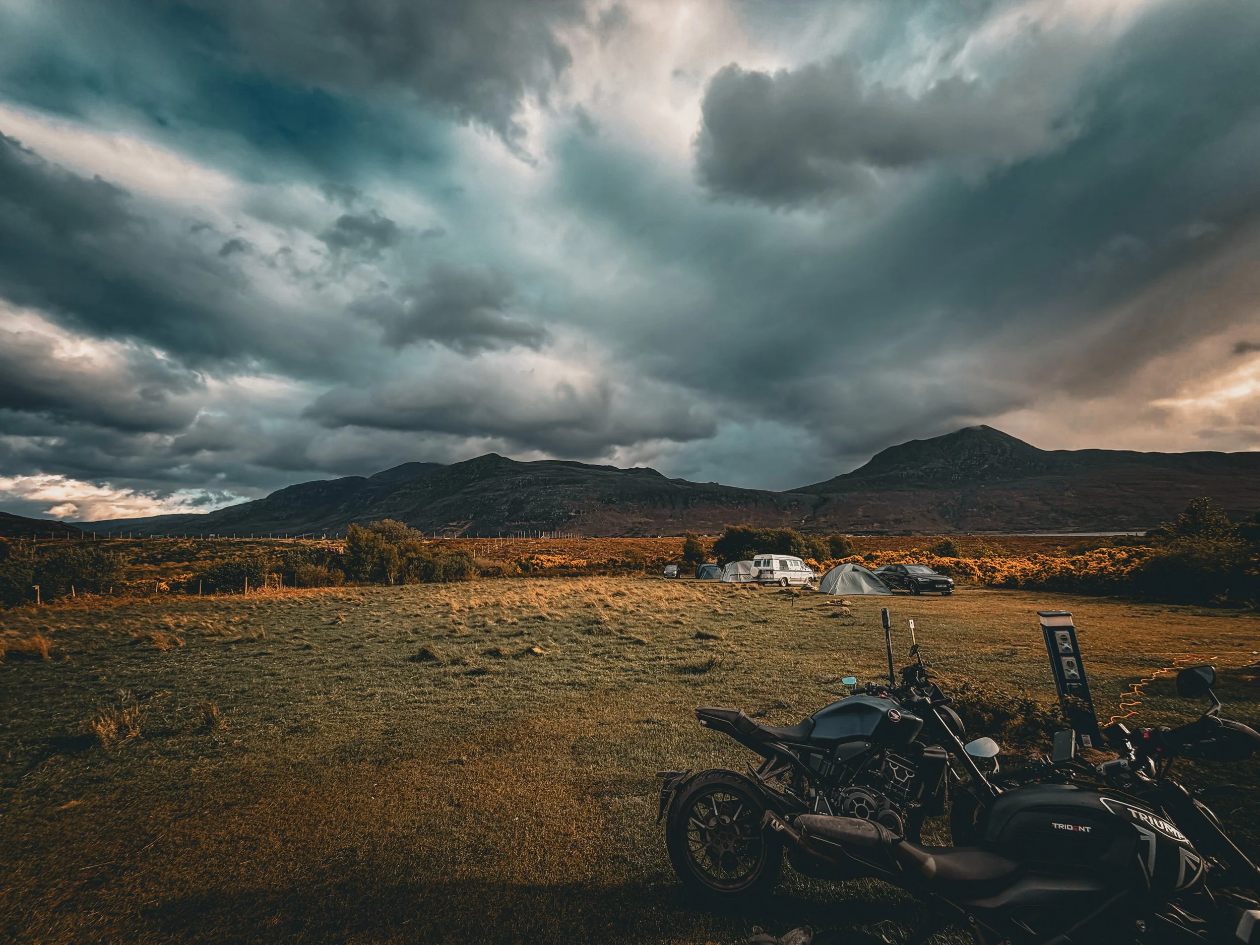 A motorcycle parked on a grassy area at a campsite with tents and vehicles, mountain range in the background, dark stormy clouds overhead.