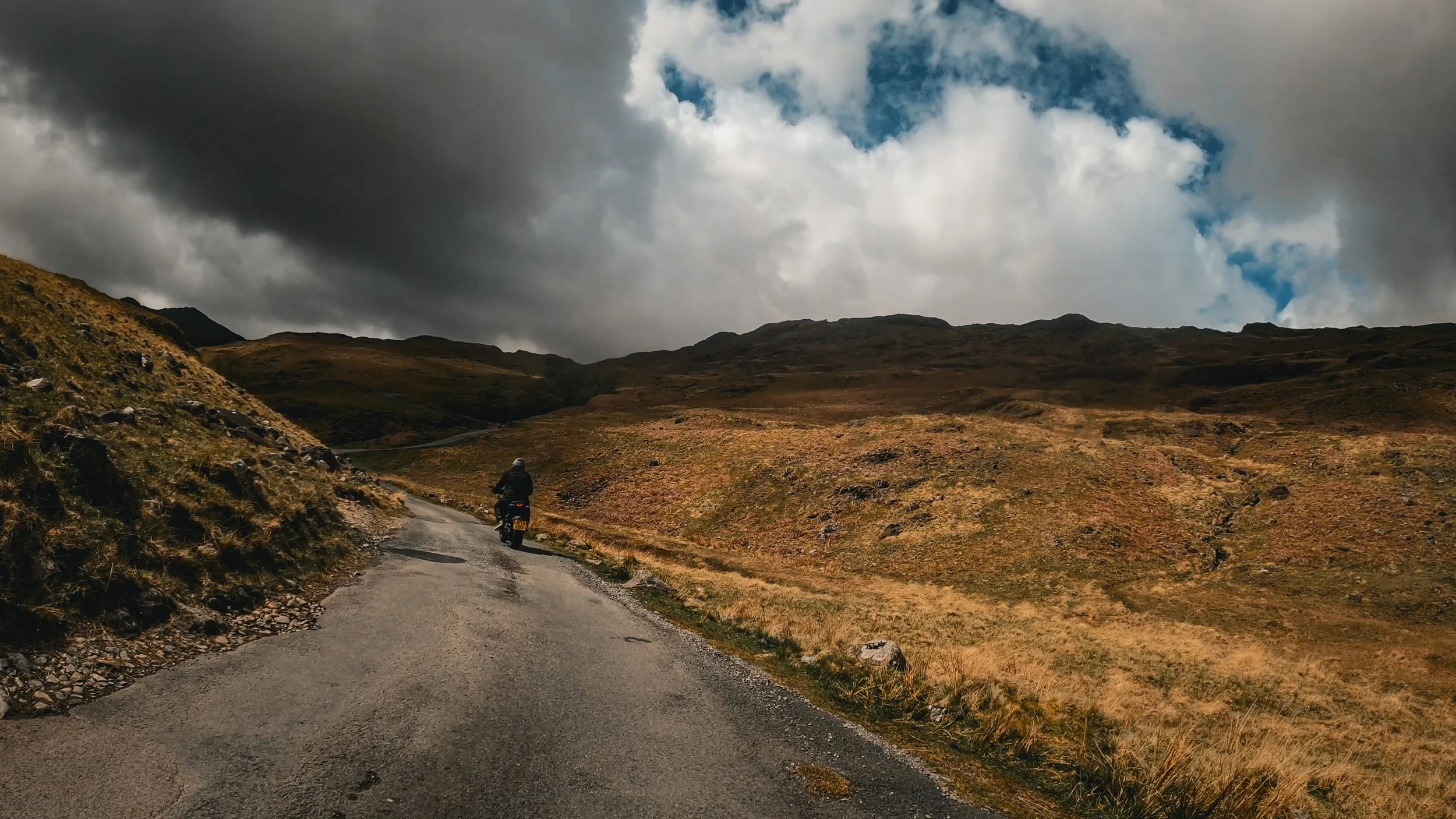 A person riding a motorcycle on a winding dirt road through a hilly, grassy landscape under a cloudy sky.