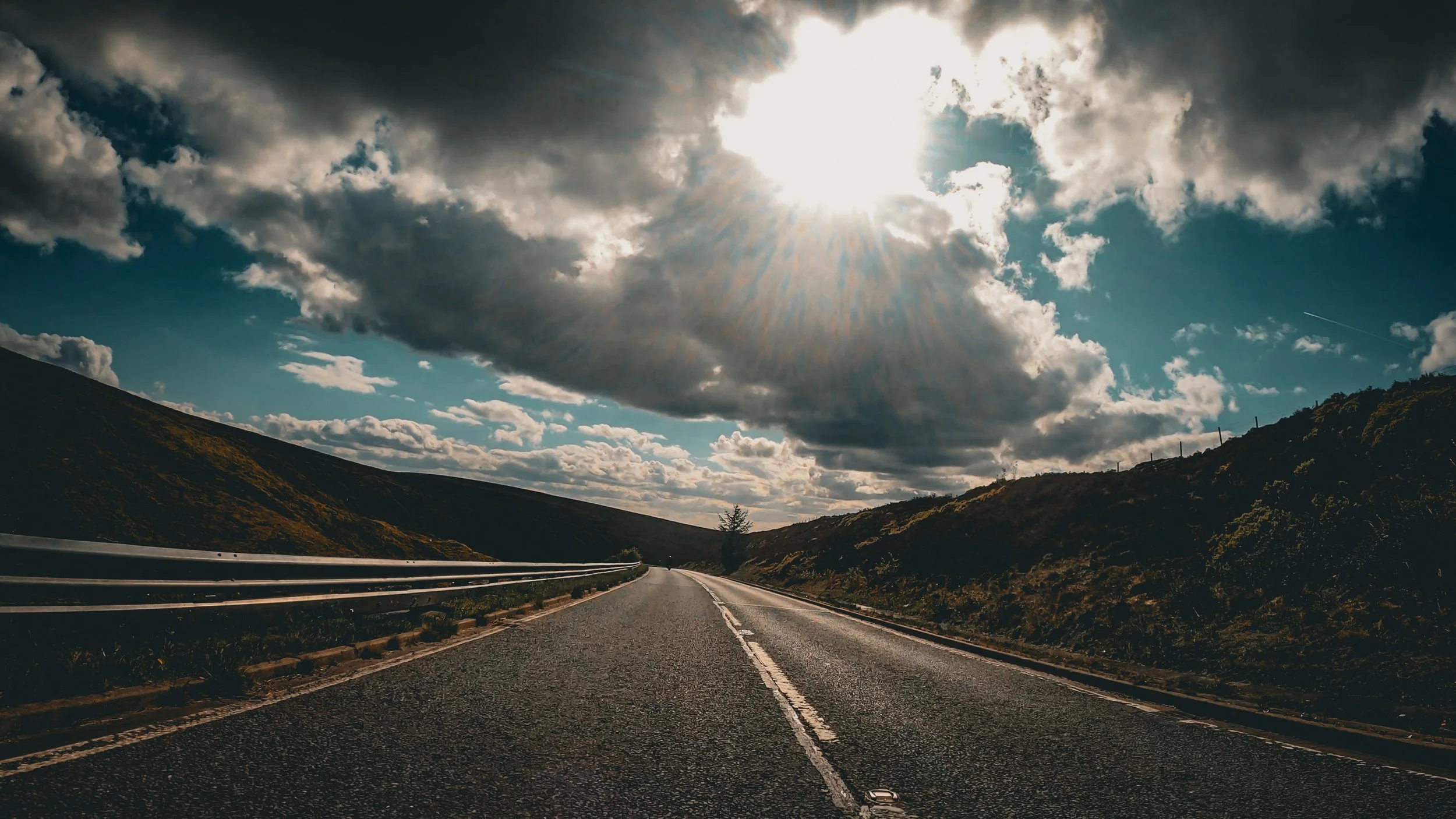 A scenic mountain road under partly cloudy skies with sun rays breaking through clouds, and a guardrail along the road.