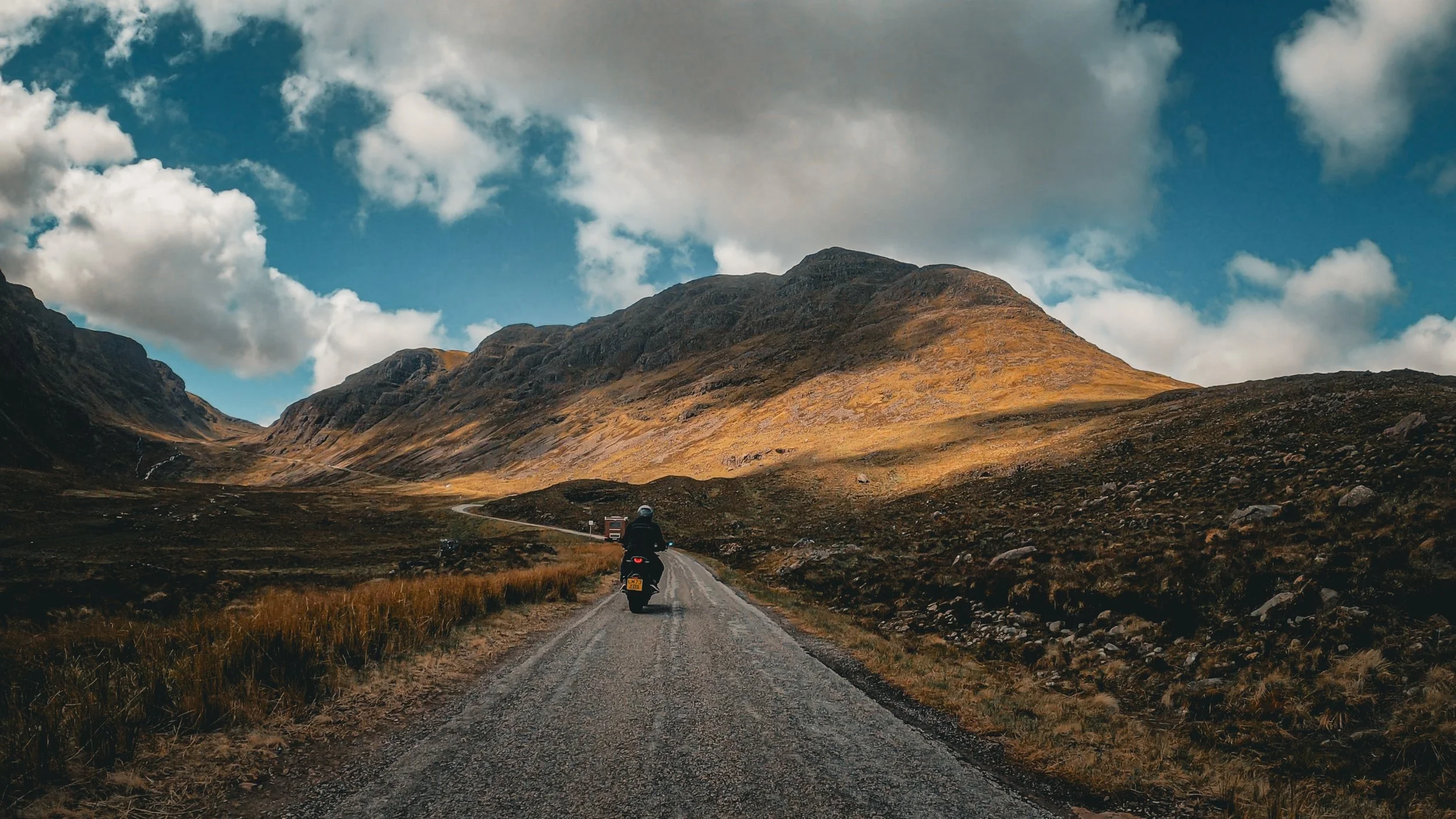 Motorcycle riding on a gravel road through a mountainous landscape with cloudy sky.