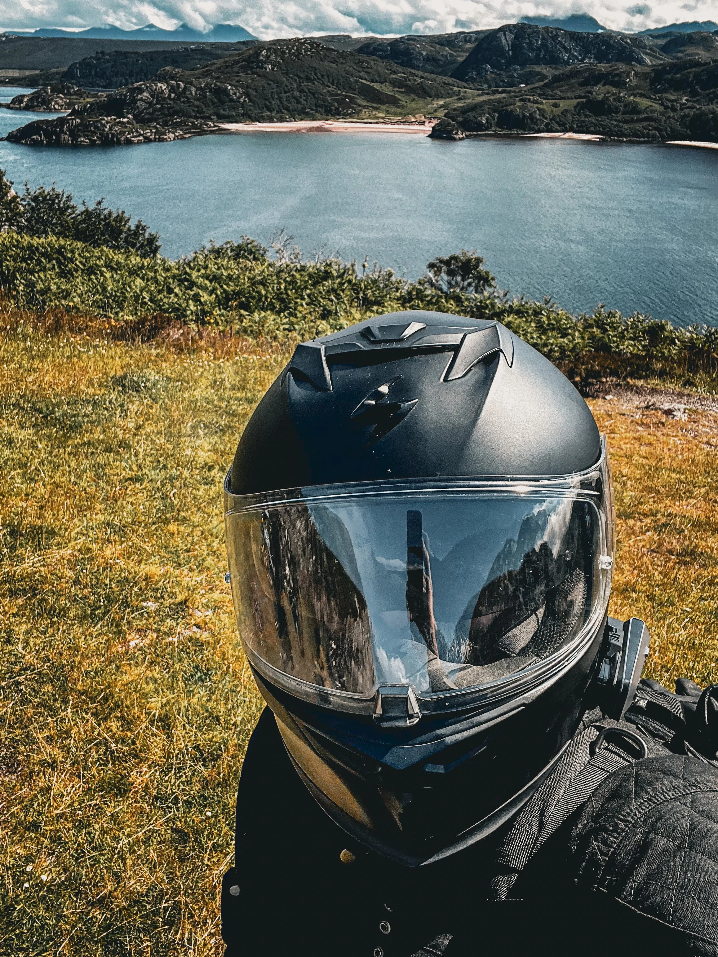 A motorcycle helmet with a reflective visor is placed on a grassy hillside overlooking a large body of water and distant mountains under a partly cloudy sky.