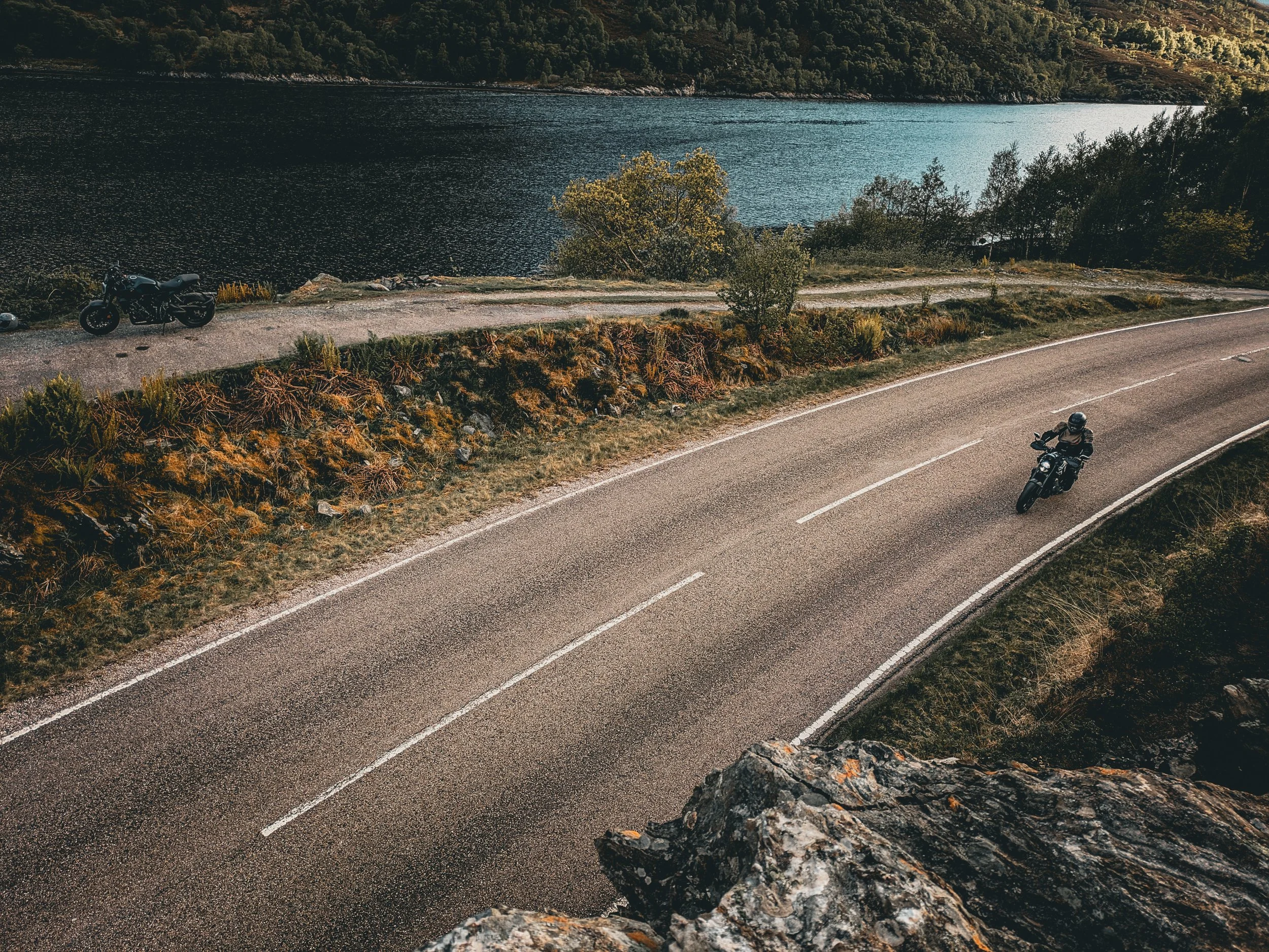 Two motorcyclists riding on a winding mountain road above a lake with forested hills in the background.