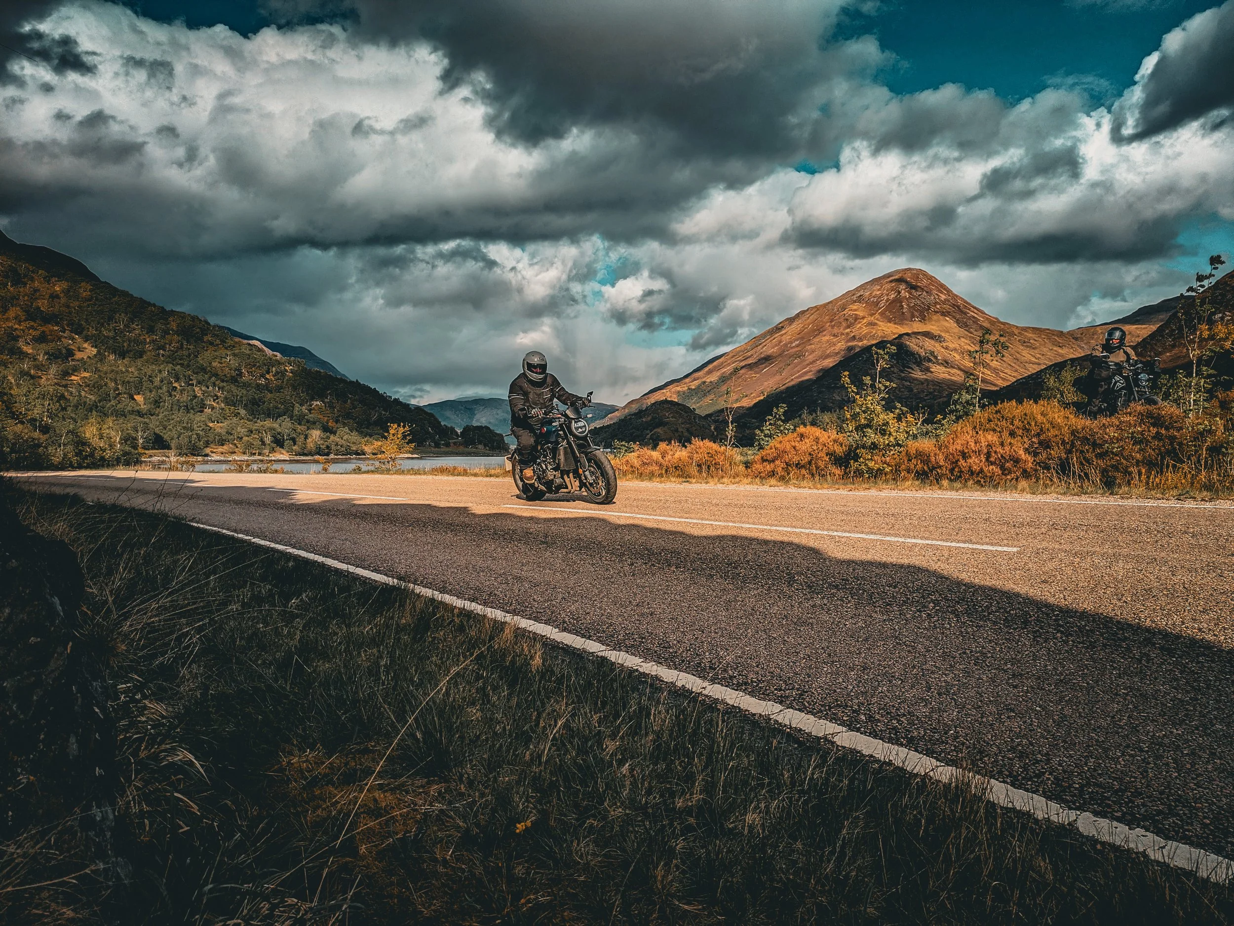 Two motorcyclists riding on a scenic mountain highway surrounded by green foliage and dramatic cloudy sky.
