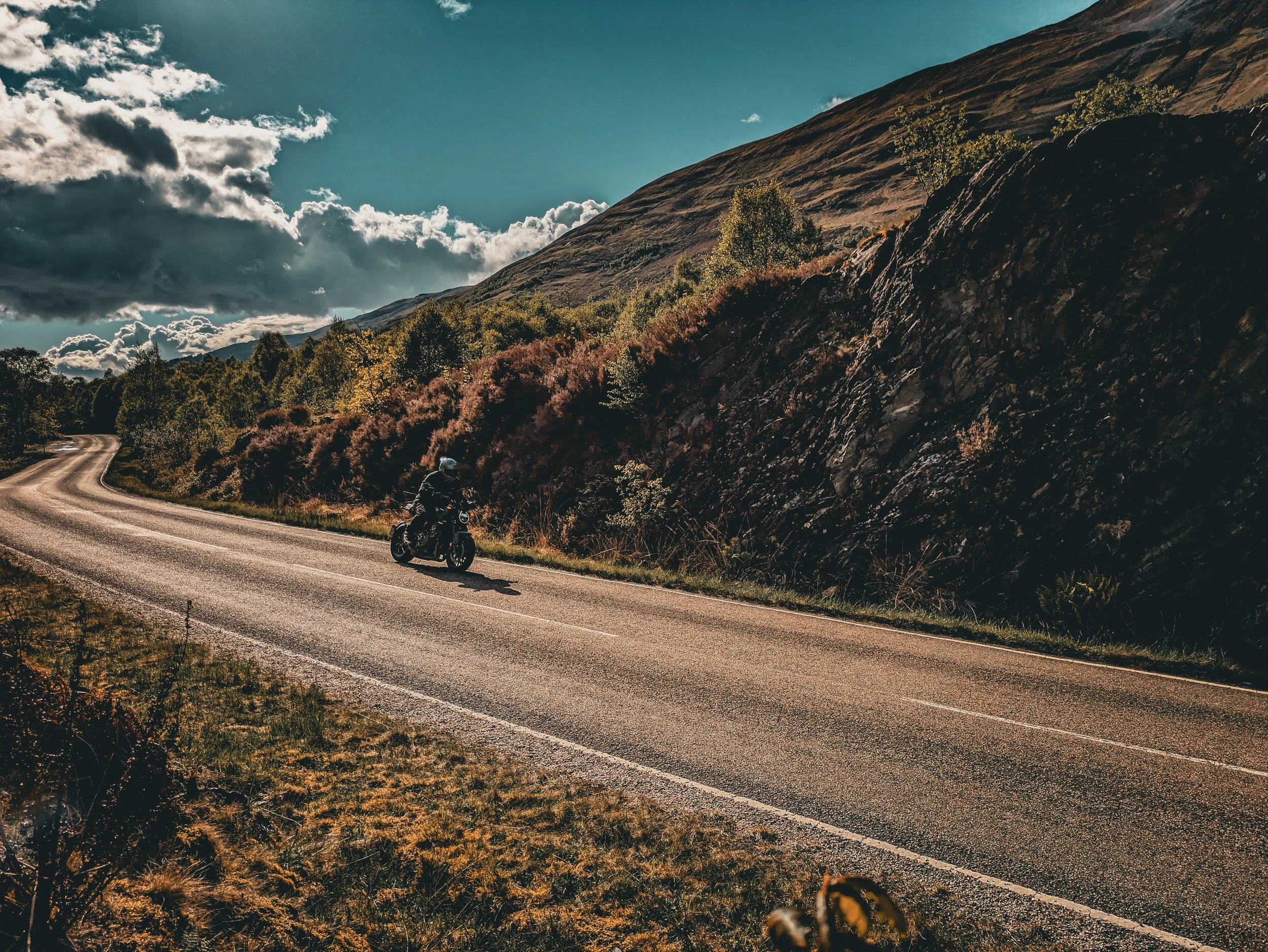 A motorcyclist riding on a winding mountain road surrounded by trees, rocky hillside, and a sky with clouds.