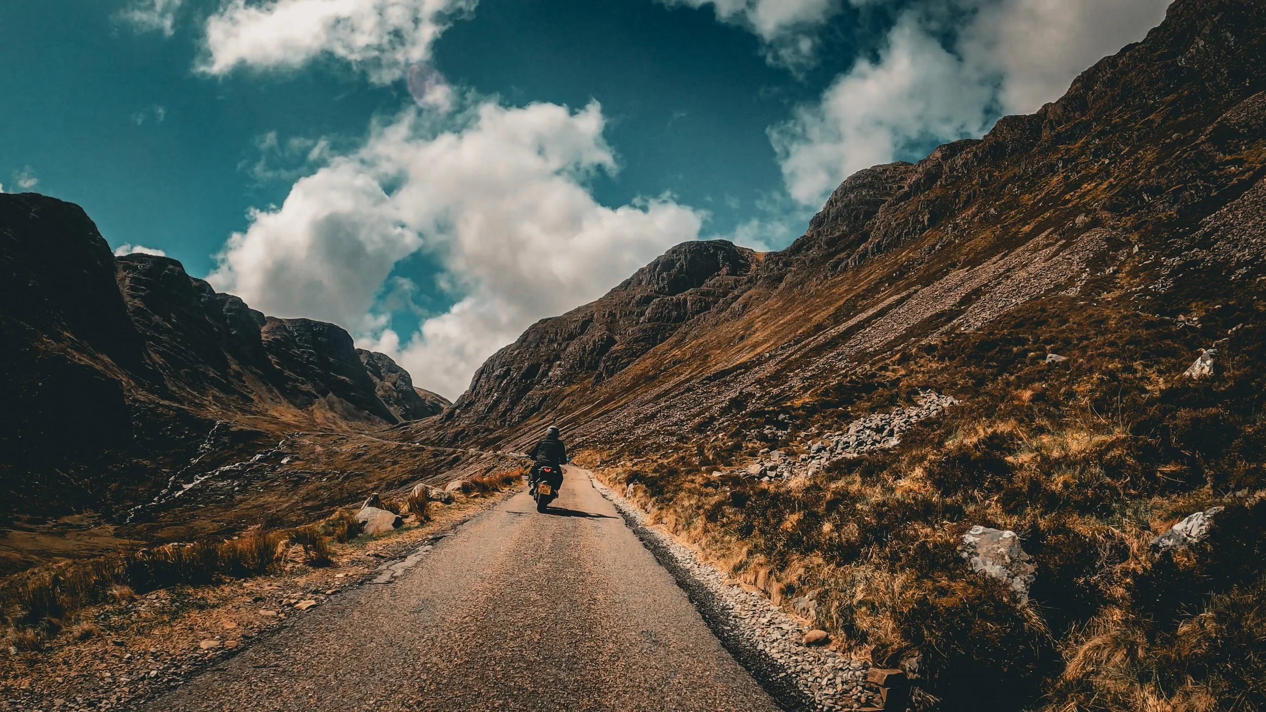 A person riding a motorcycle on a narrow mountain road surrounded by rocky hills and mountains under a partly cloudy sky.