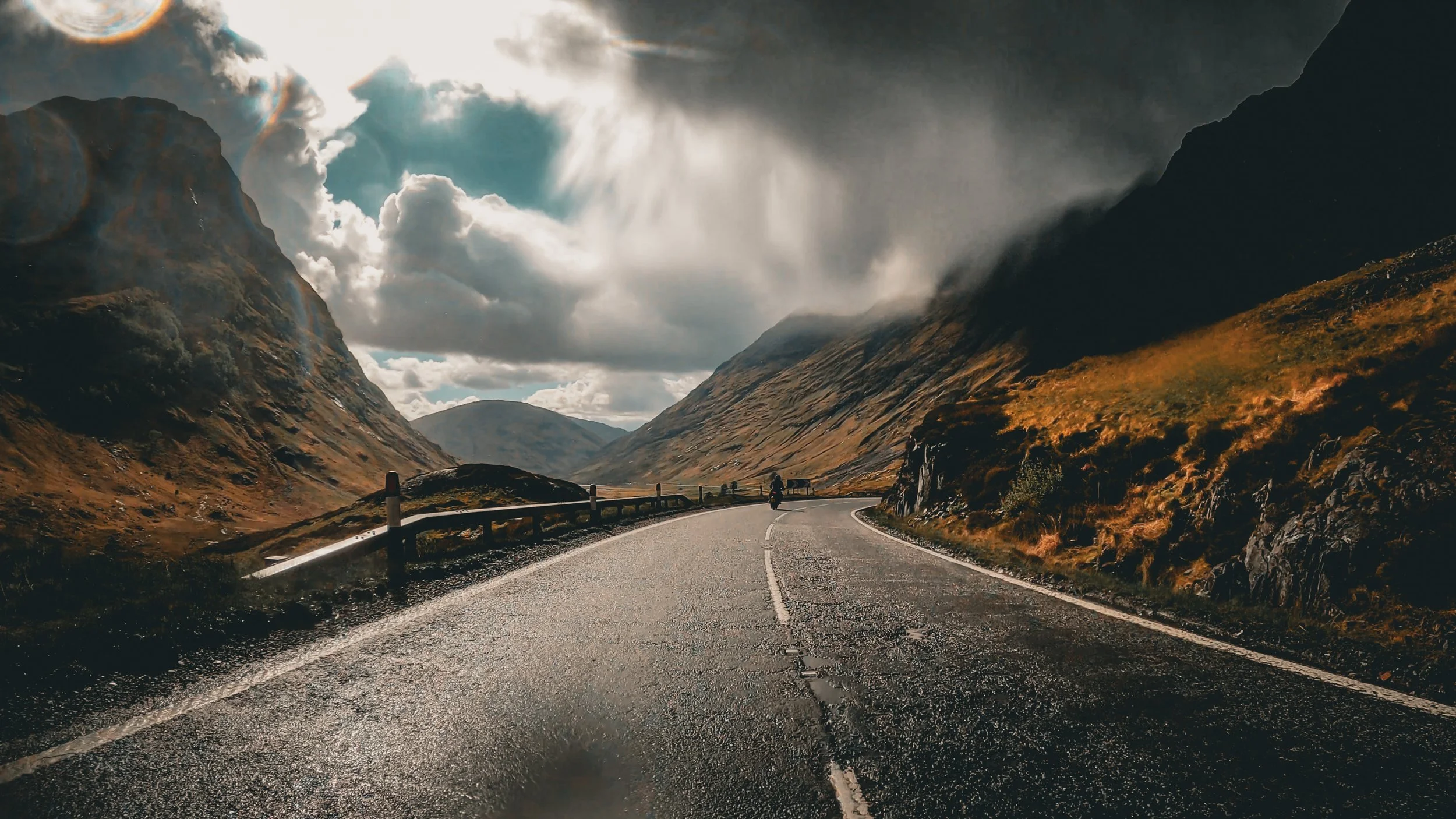 A scenic mountain road winding through a valley with tall, rocky mountains on either side, partly cloudy sky with sunlight breaking through, and a motorcyclist riding along the road.