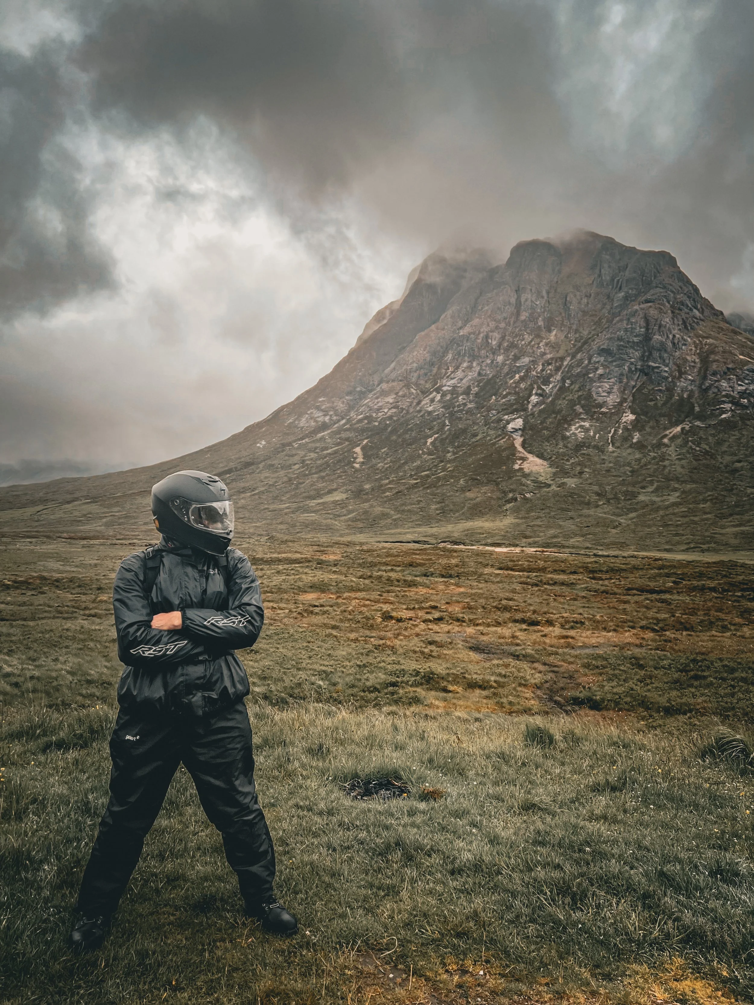 A person in black gear and a helmet standing outdoors on a grassy field with a mountain and cloudy sky in the background.