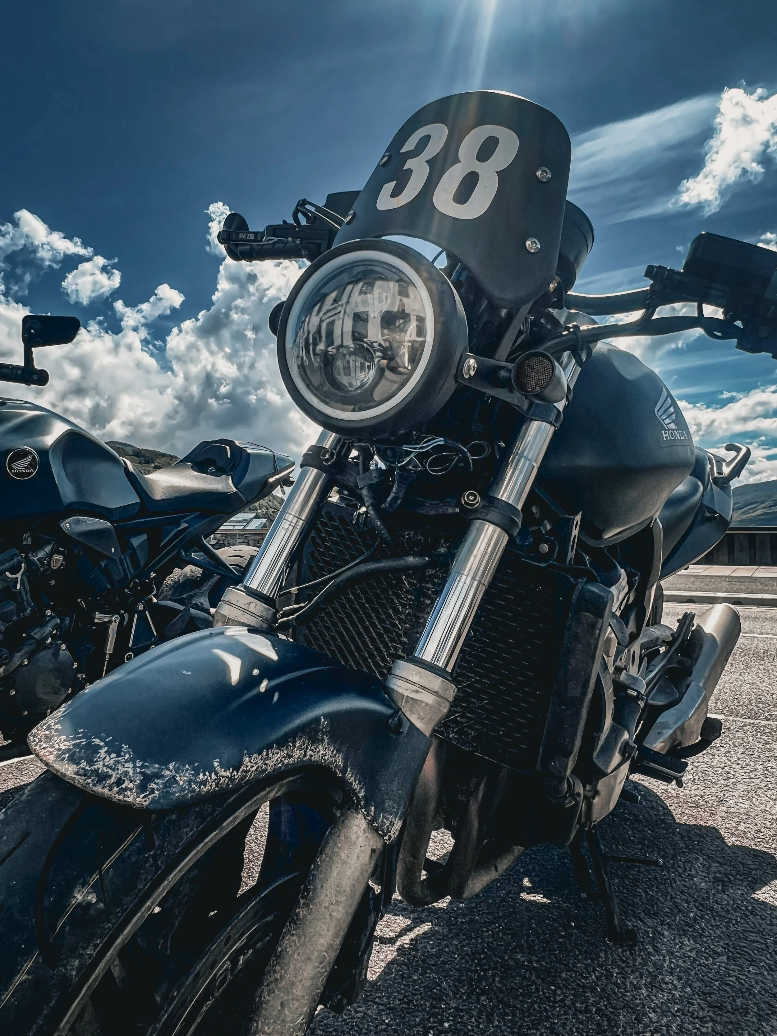 Close-up of a black Honda motorcycle with a cloud and blue sky background, front view showing the headlight, front fender, and the number 38 on the windscreen.