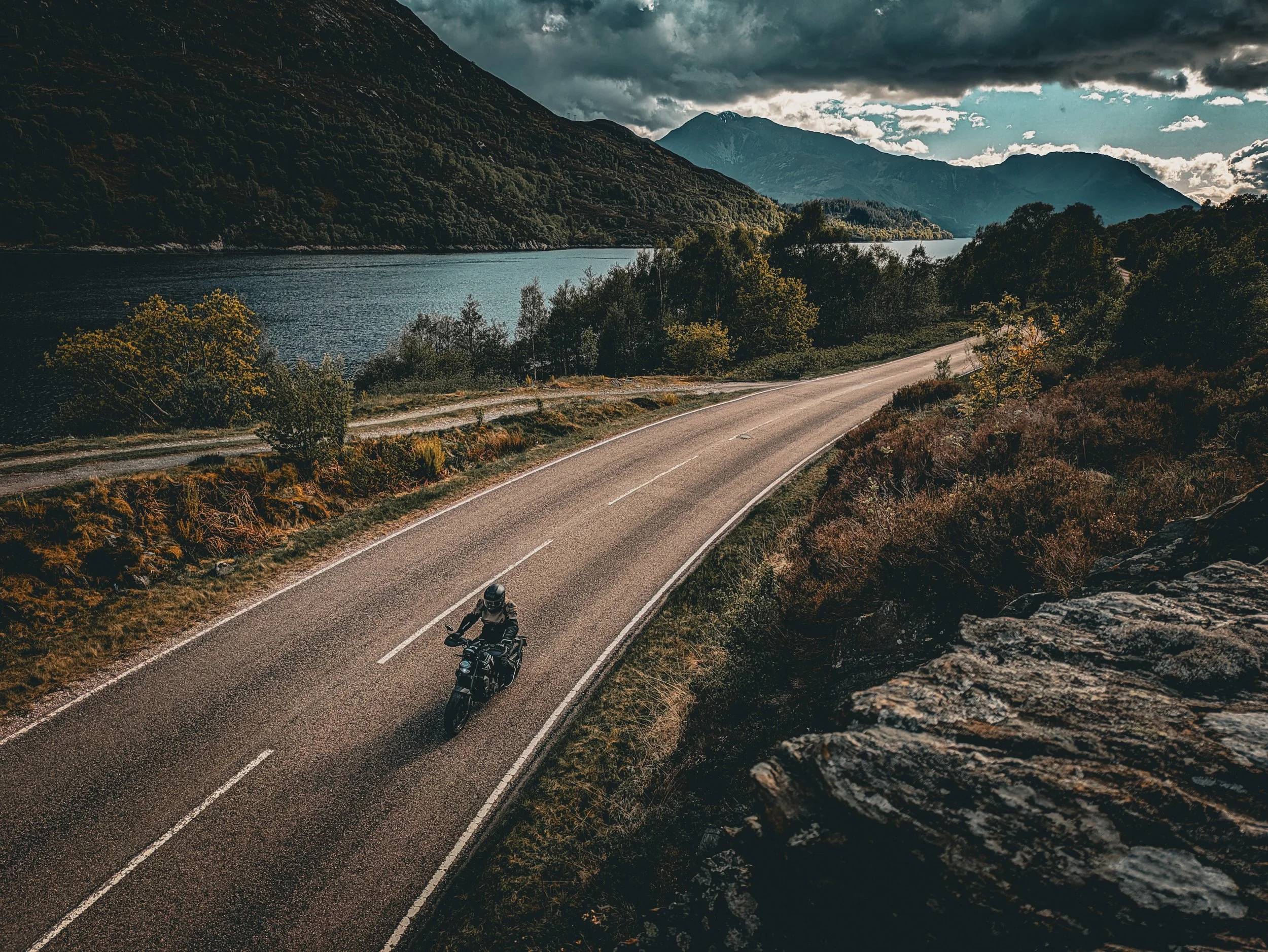 Motorcycle riding along a winding mountain road by a body of water, with forested hills and mountains in the background under a cloudy sky.
