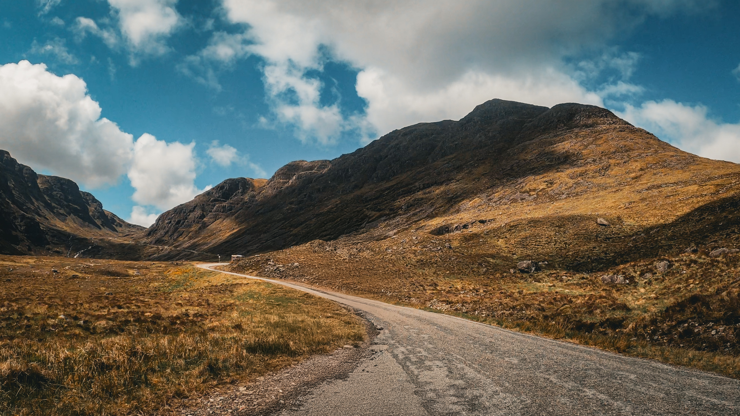 A winding dirt road through a valley with brown and green grass, flanked by mountains under a partly cloudy sky.