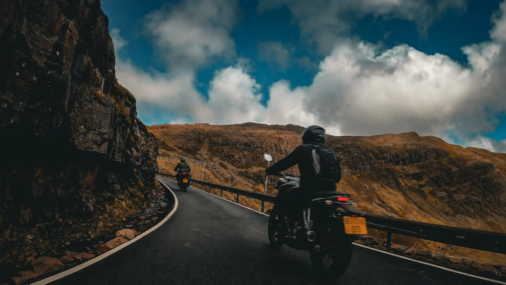 Two motorcyclists riding along a winding mountain road with rocky hills and a partly cloudy sky in the background.