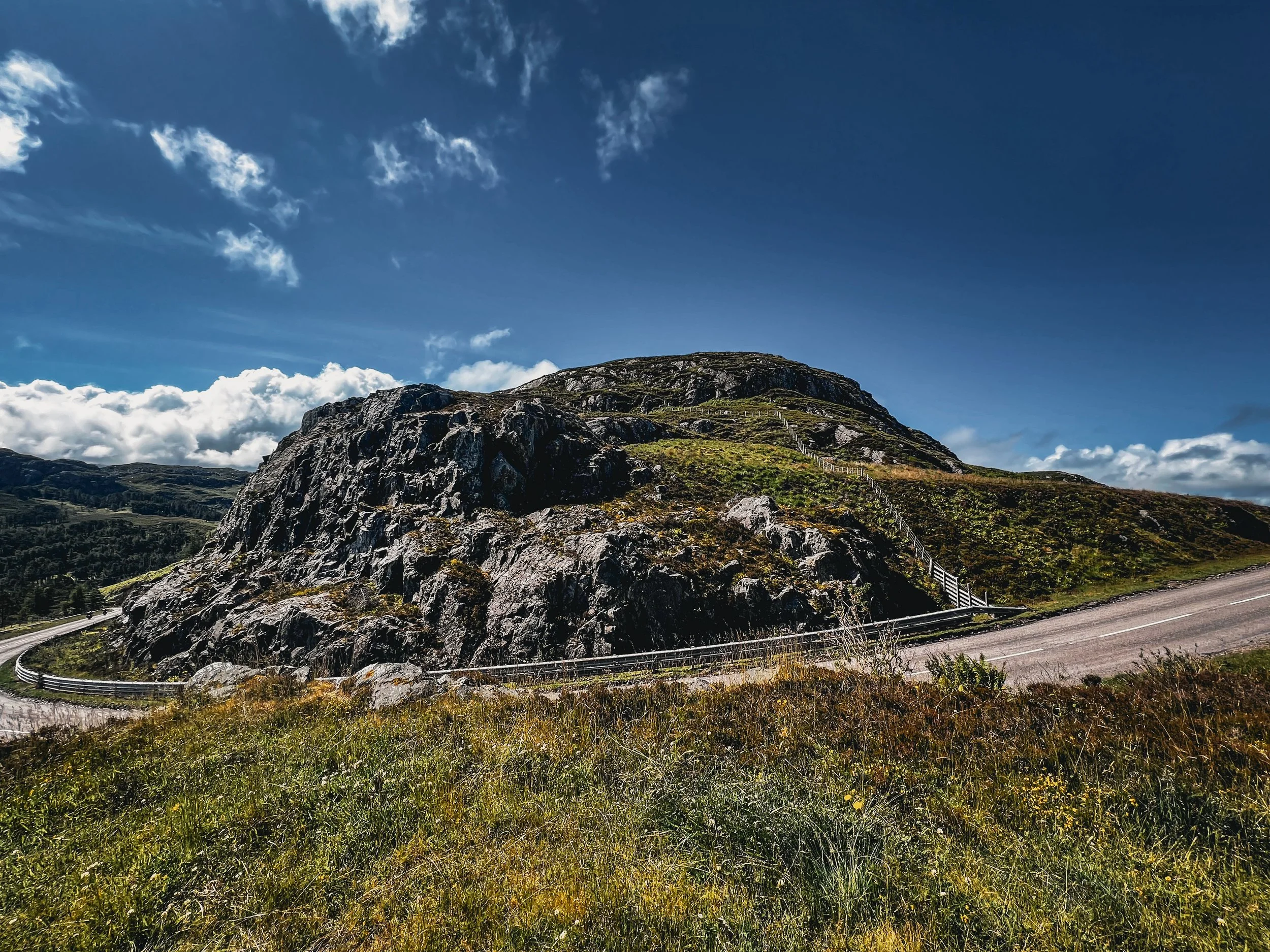 A mountain with a winding road and a cable fence on the hillside under a partly cloudy sky.