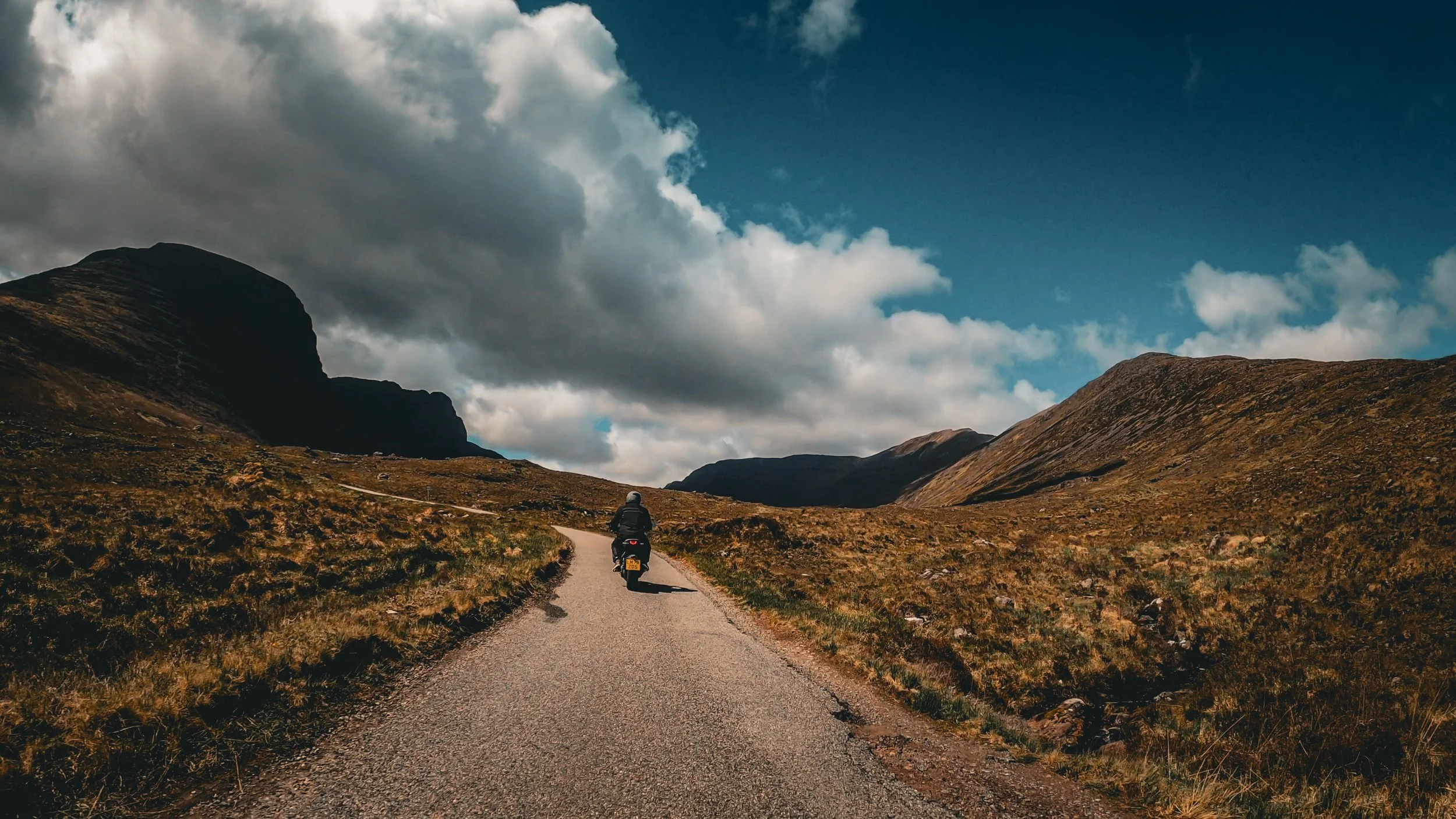 Motorcyclist riding on a winding road through mountainous terrain under a partly cloudy sky.
