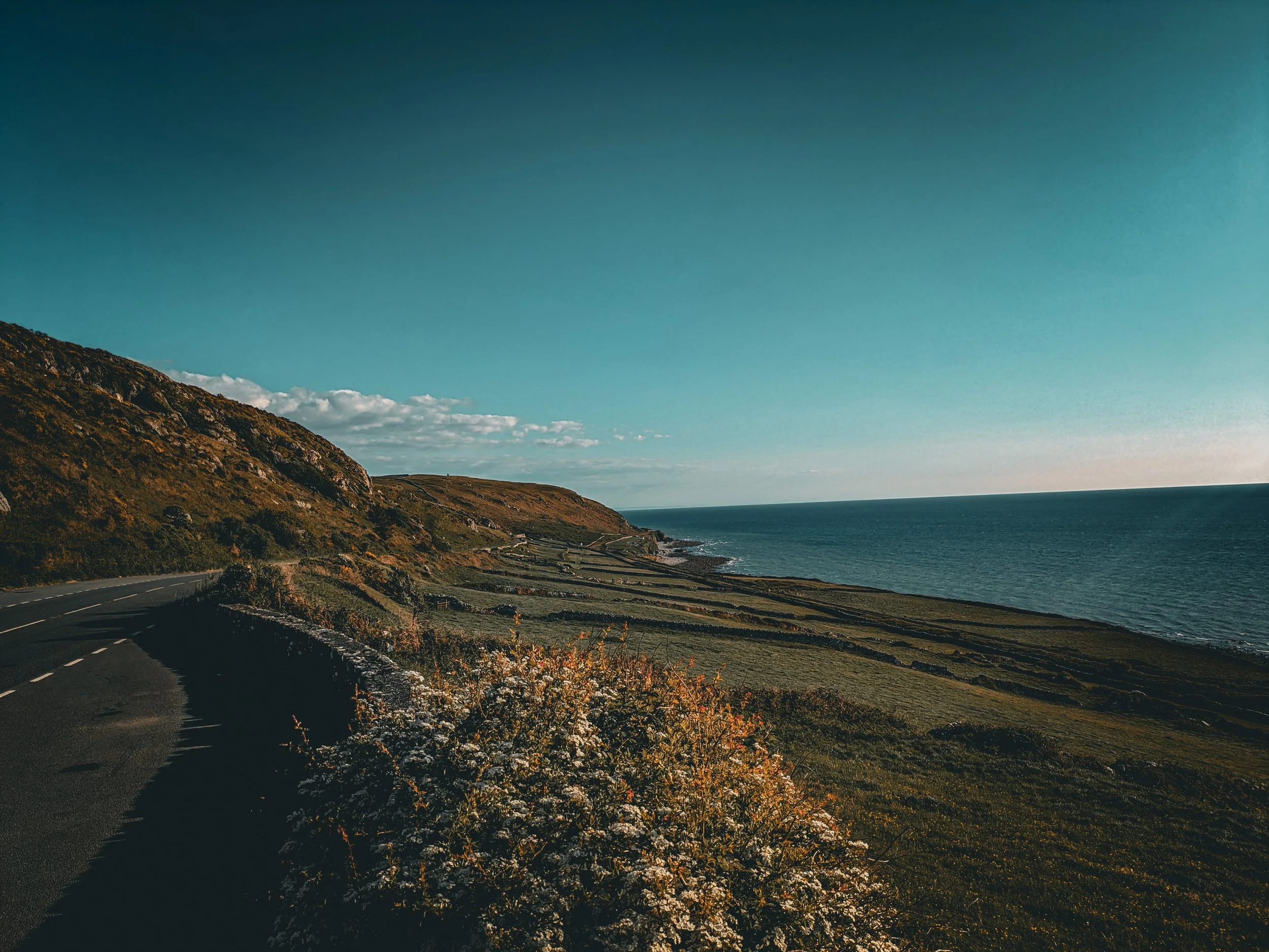 Scenic view of a coastal highway along the rugged hillside with the ocean on the right, under a clear blue sky with a few clouds.