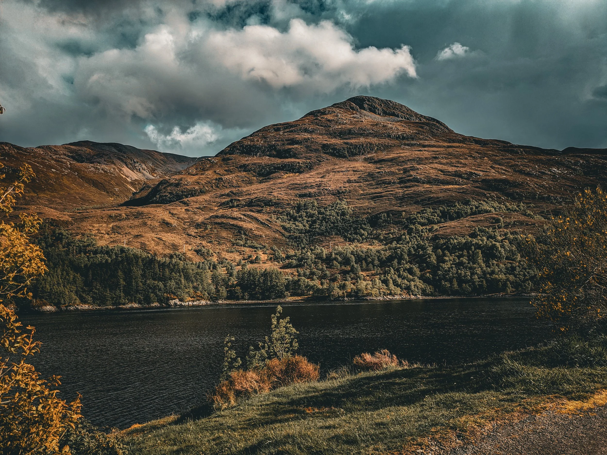 A scenic view of a mountain landscape with a lake at the base and cloudy sky overhead, showing rolling hills and trees.