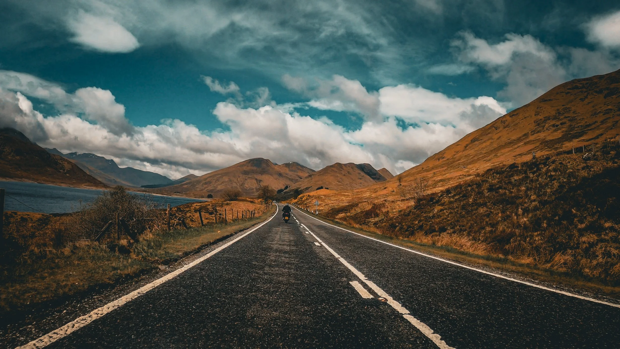 A motorcycle traveling on a two-lane road through a mountainous landscape with clouds in the sky.