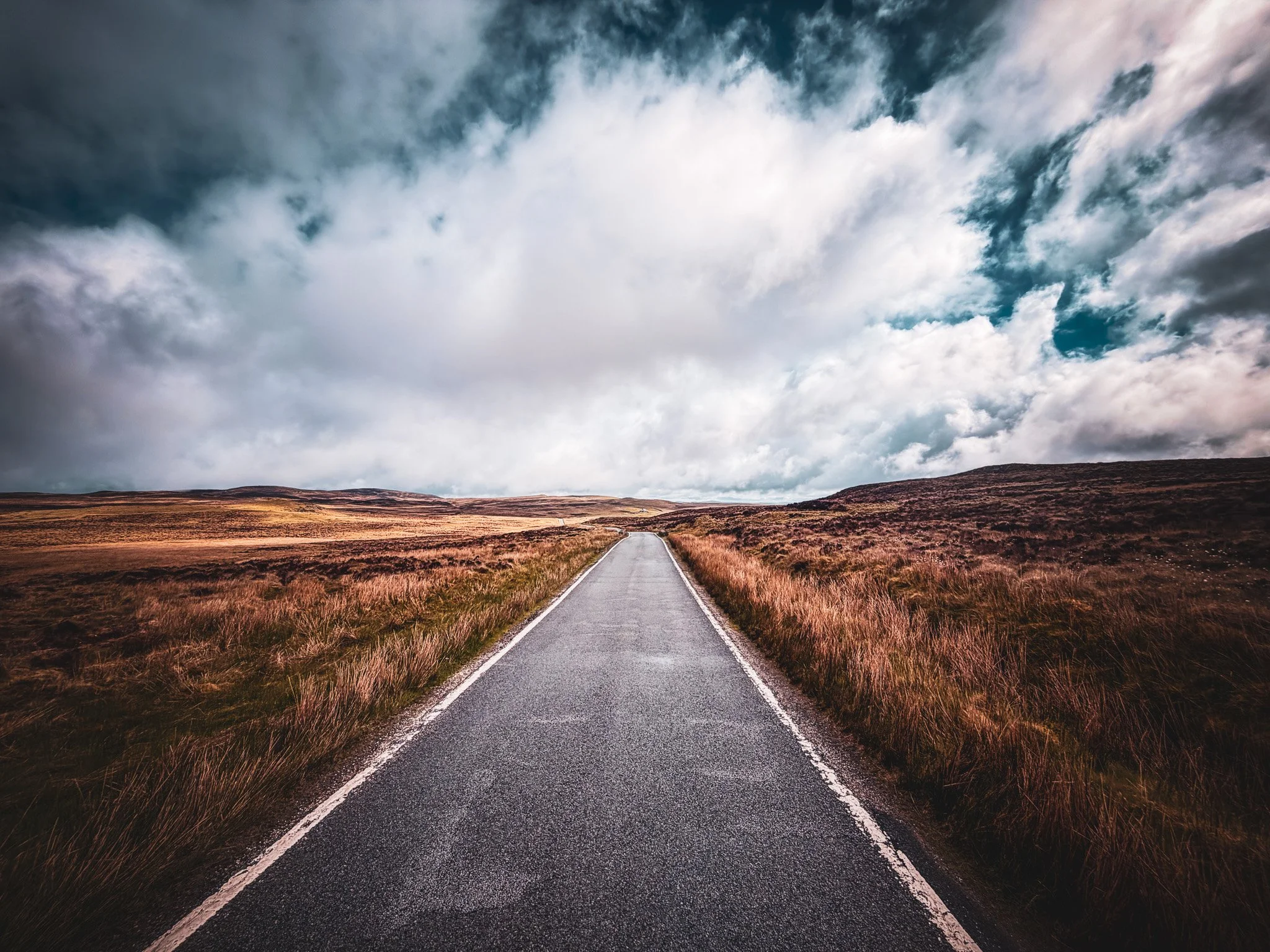A straight paved road extending into the horizon through open countryside with brownish grass on either side, under a partly cloudy sky with dark and light clouds.