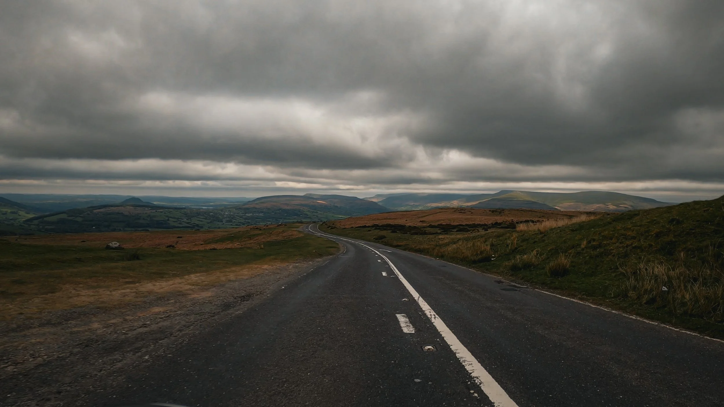 A winding two-lane road runs through a green hilly landscape under a cloudy gray sky.