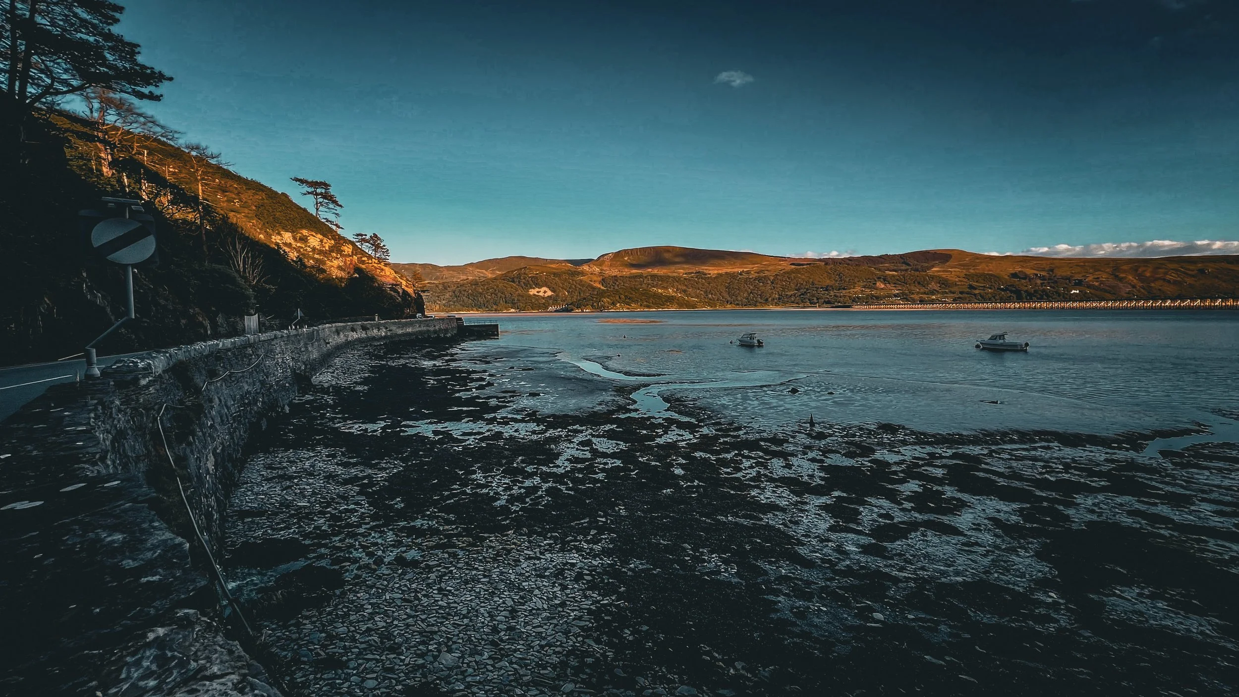 Coastal scene with a rocky shoreline, two boats in the water, green hills in the distance, and a blue sky with some clouds.