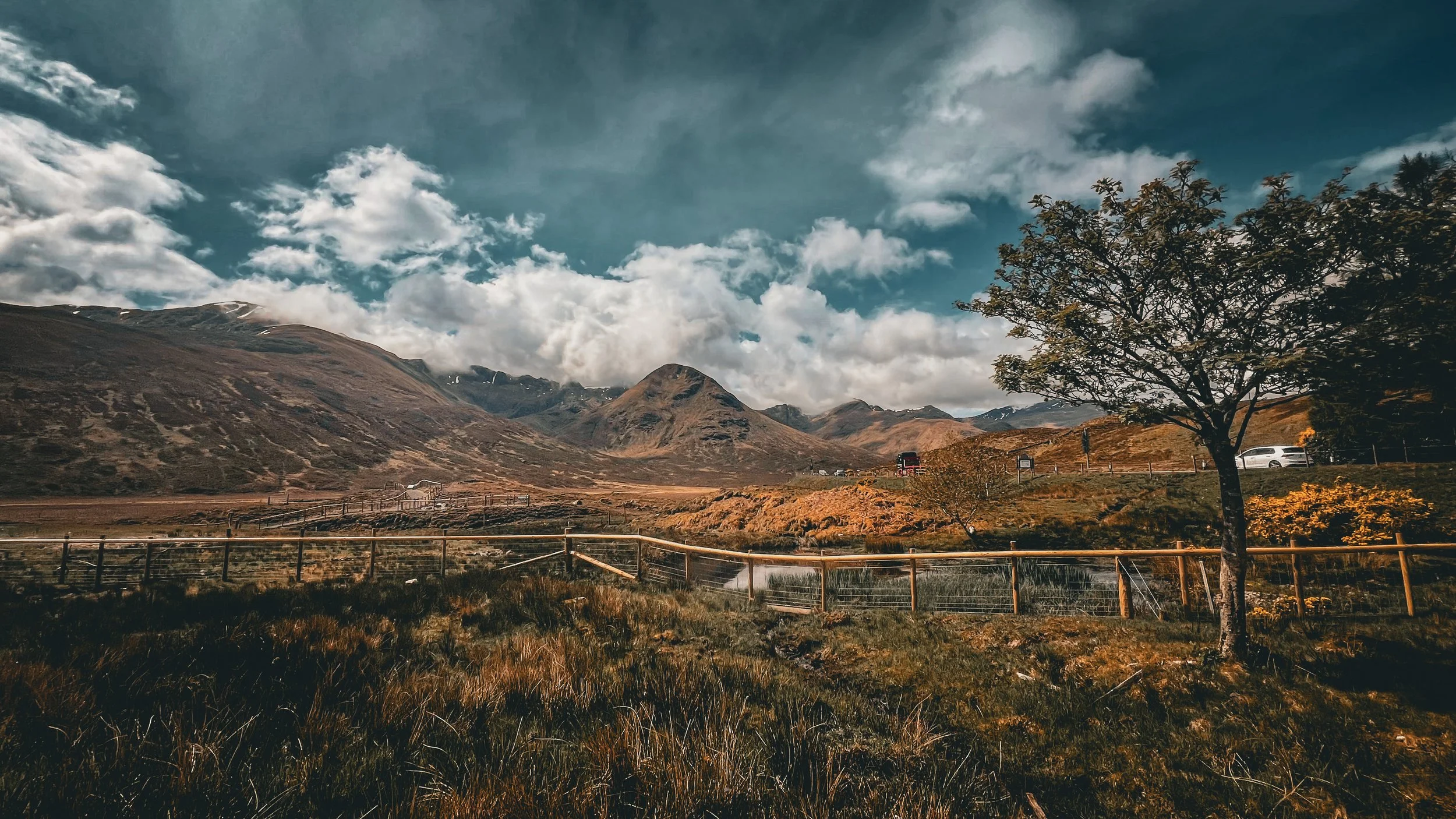 Scenic landscape of mountains, cloudy sky, and a tree in a fenced area with vehicles and a small building in the distance.