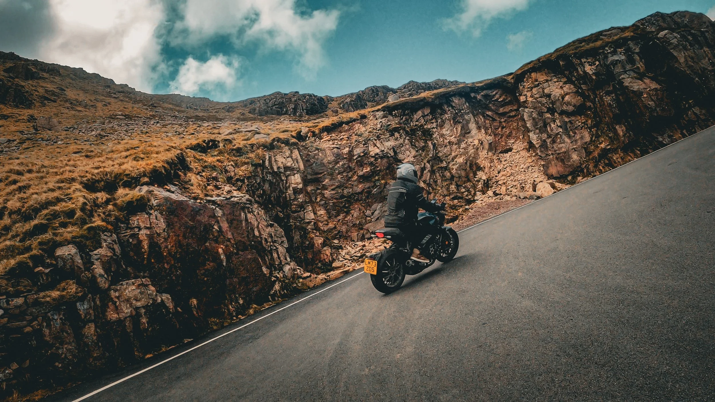 Motorcycle rider wearing a helmet and riding on a winding mountain road with rocky, grassy terrain on the side and a blue sky with clouds above.