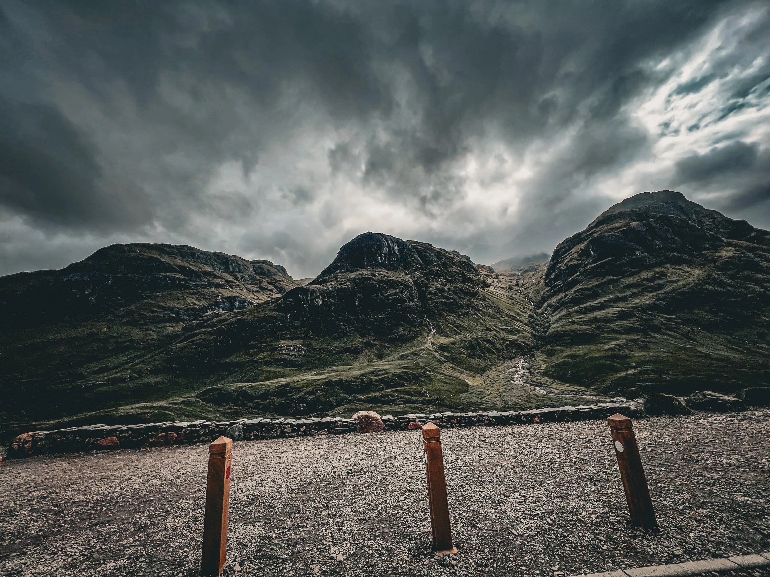 A mountainous landscape under a cloudy sky, with a gravel parking area in the foreground bordered by wooden posts.