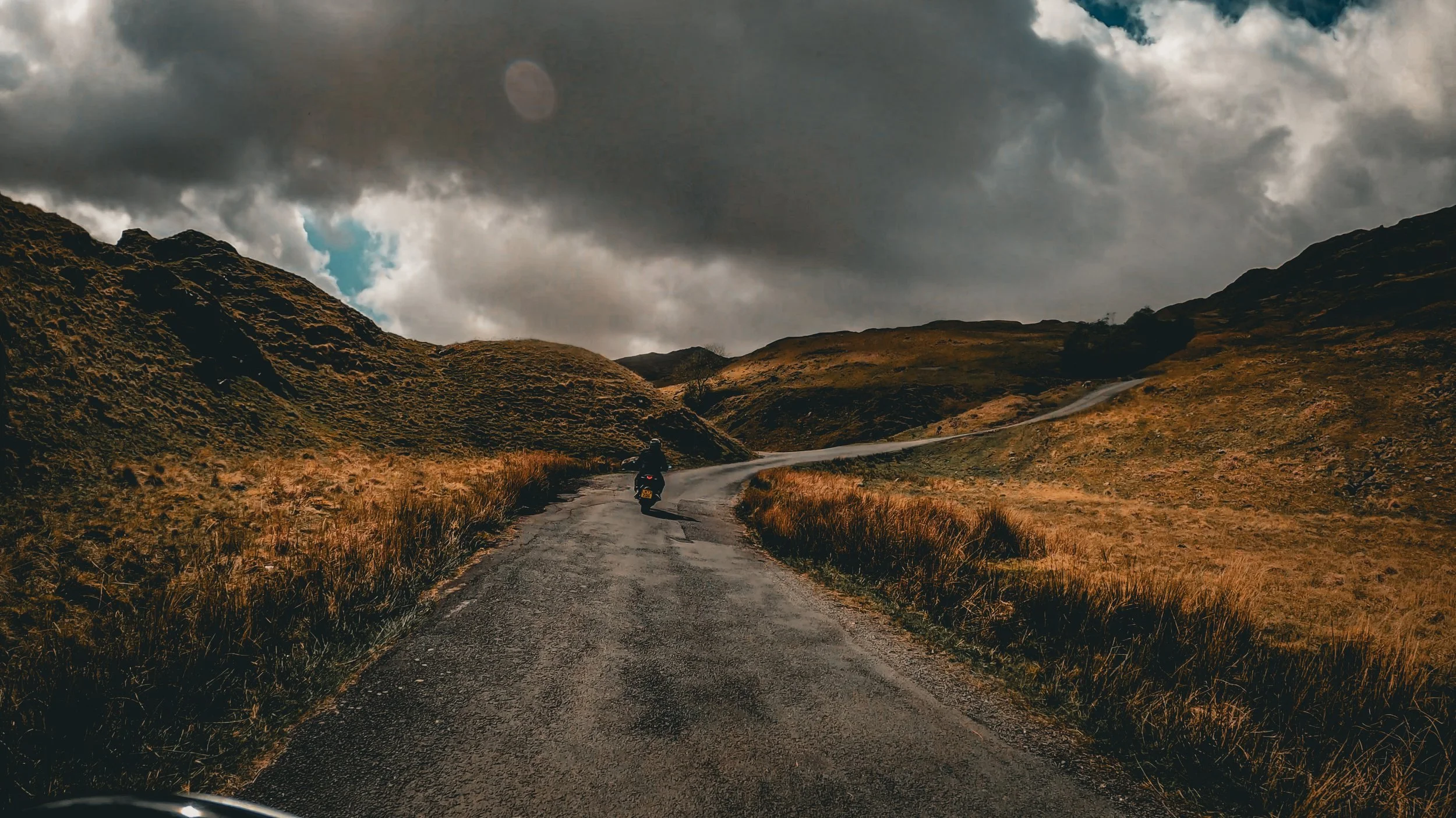A person riding a motorcycle on a winding dirt road through hilly terrain under a cloudy sky.