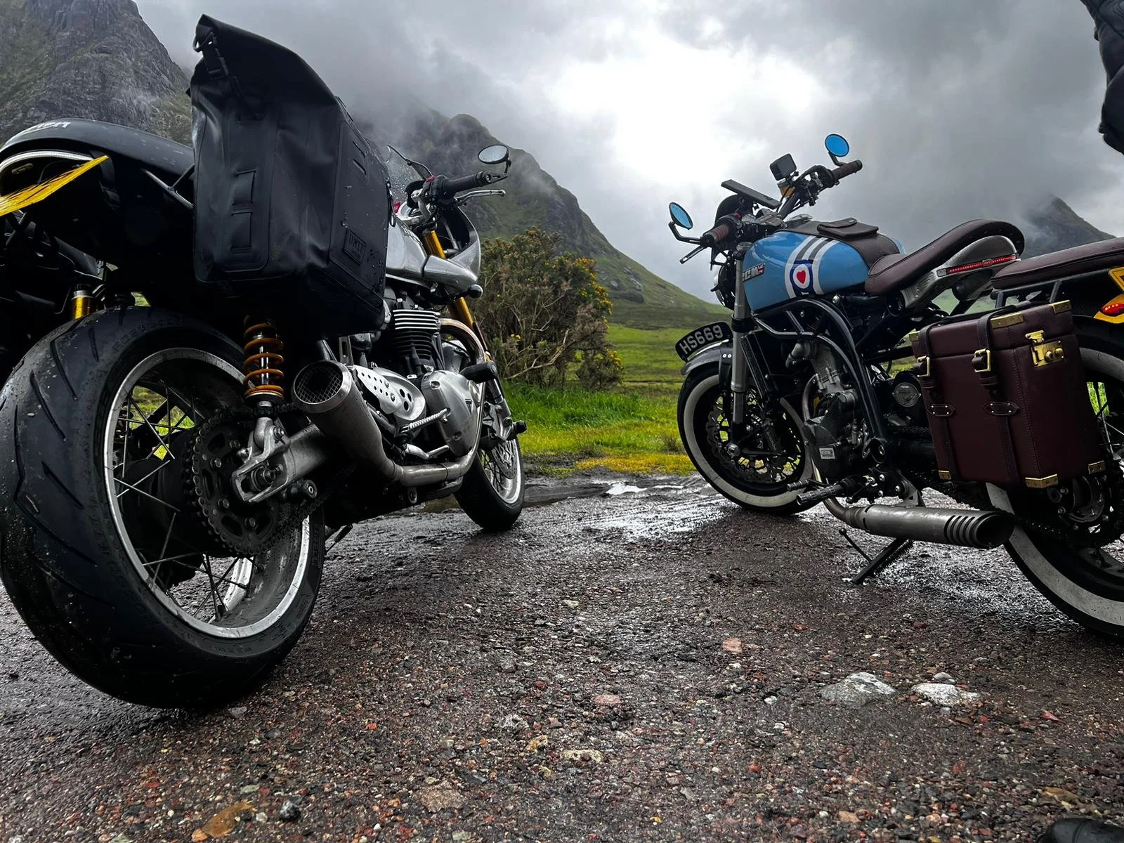 Two motorcycle bikes parked on a wet, muddy ground with a lush green landscape and mountains in the background under a cloudy sky.