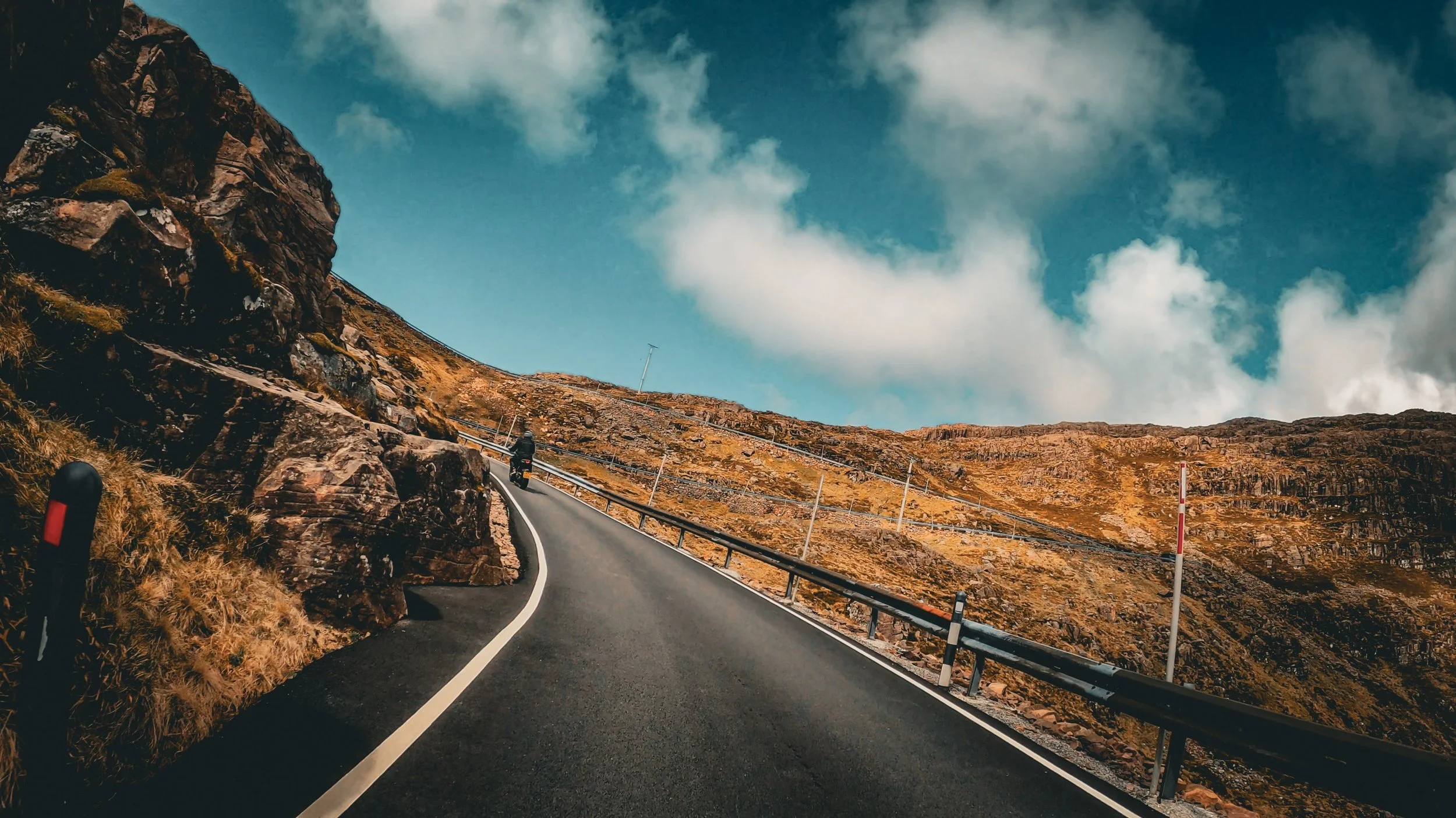 Motorcycle traveling on a winding mountain road with rocky cliffs and a partly cloudy sky.