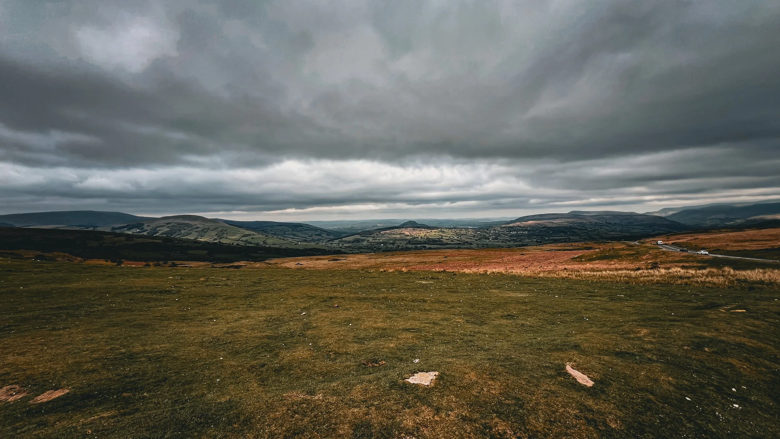 A landscape scene with rolling hills under a dark, cloudy sky. There is a grassy field in the foreground and a distant road with a few cars on it.