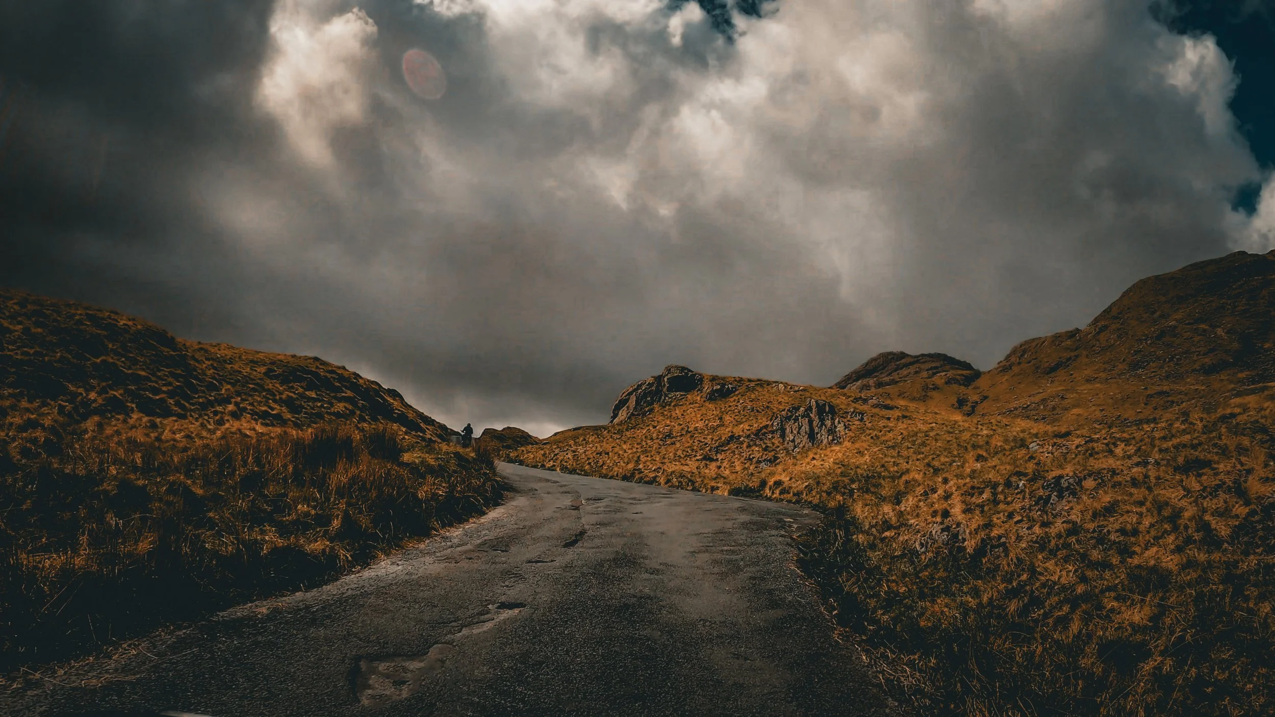 A dirt road winds through a mountainous landscape with grassy hills and rocky outcroppings under a dark, cloudy sky.