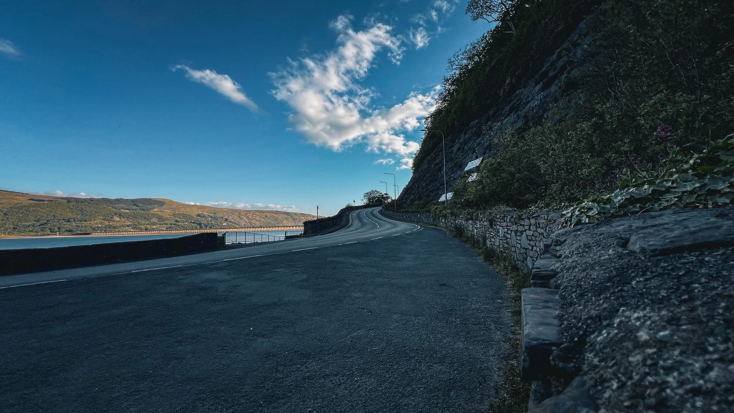 A winding mountain road alongside a body of water with a stone wall on one side, trees, and hills under a partly cloudy blue sky.