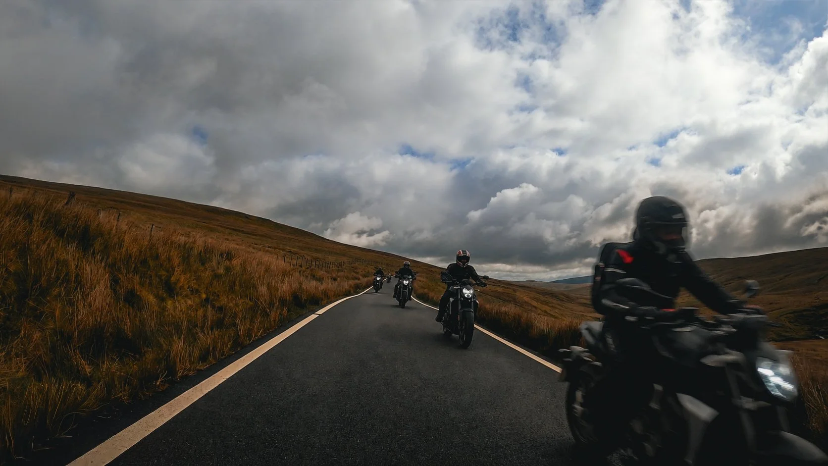 Four motorcyclists riding on a scenic, winding mountain road surrounded by grassy hills and a cloudy sky.