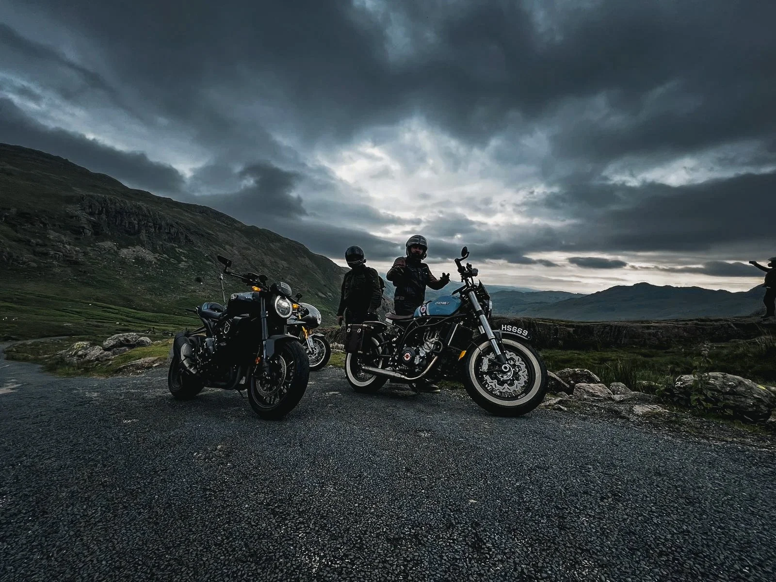 Three motorcyclists wearing helmets and riding gear standing beside their bikes on a mountain road during cloudy weather, with mountains and a cloudy sky in the background.