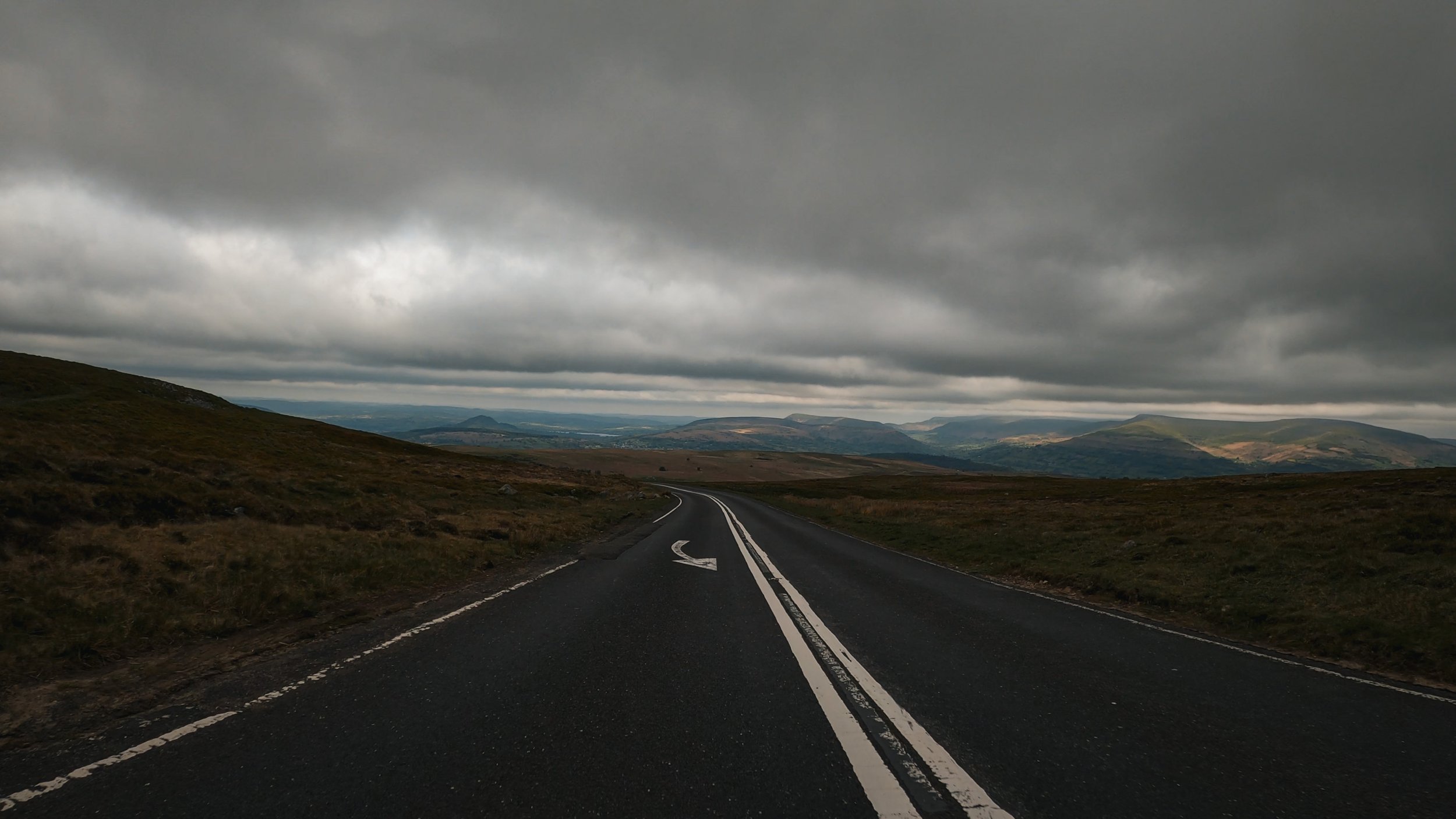 A winding road cutting through a hilly landscape under a dark, cloudy sky.