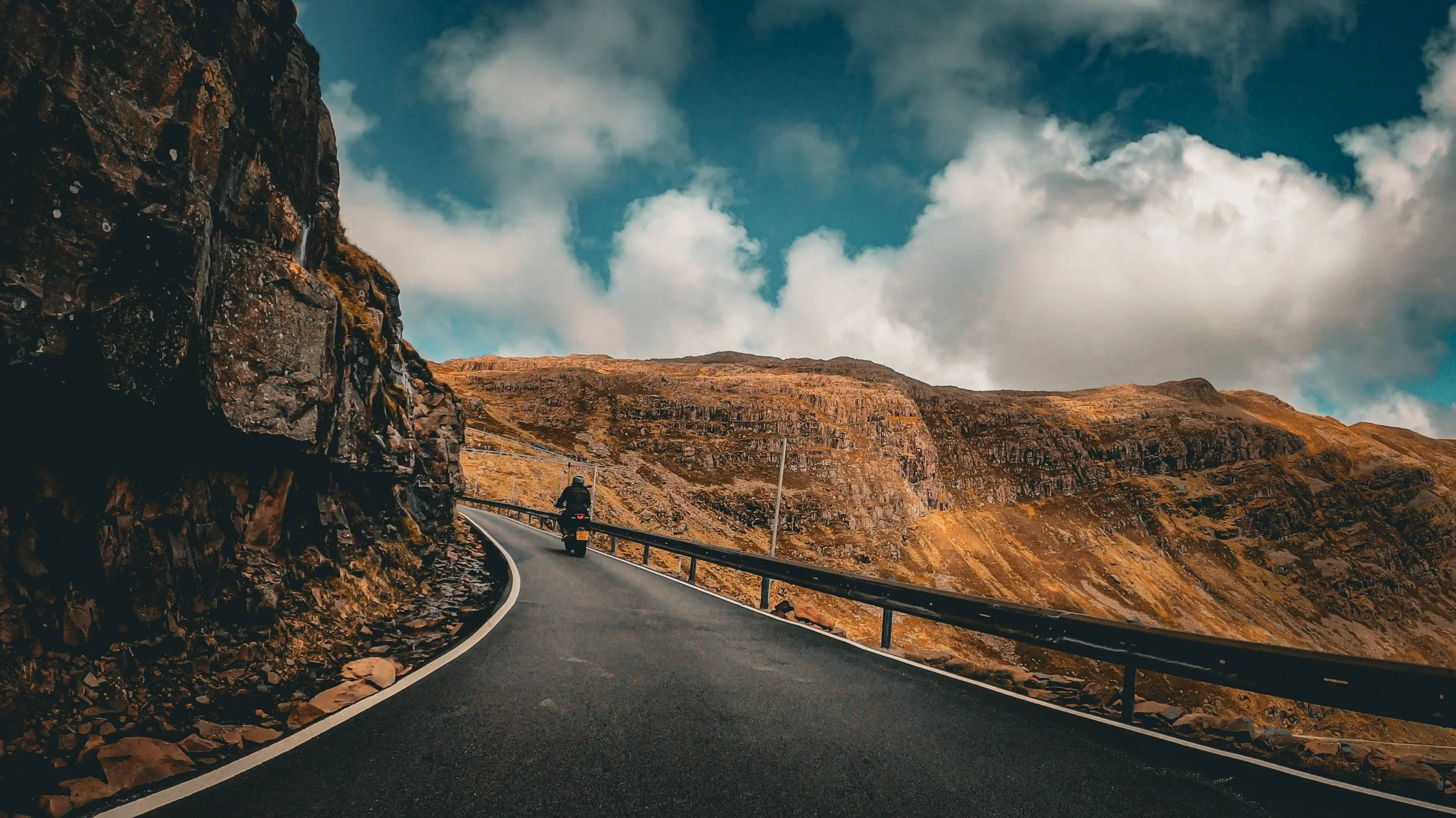 A winding mountain road with a motorcyclist riding along, surrounded by rocky hills and a partly cloudy sky.