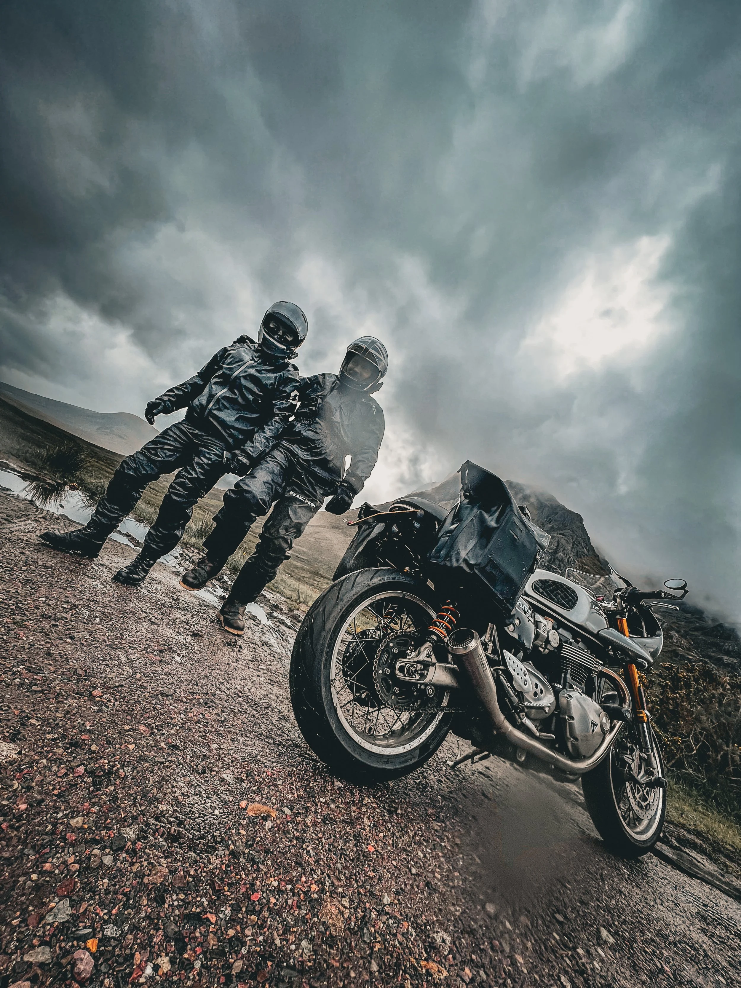 Two motorcyclists dressed in black riding gear and helmets standing next to their motorcycles on a gravelly roadside under a cloudy, stormy sky with mountains in the background.