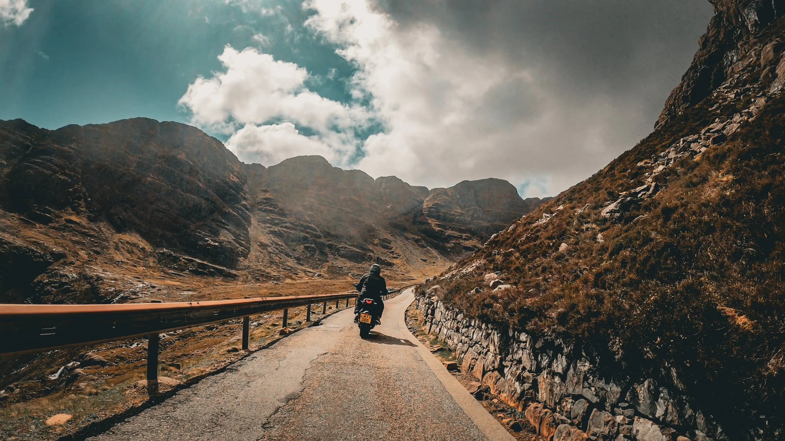 Motorcycle rider riding on a winding mountain road with rugged hills on either side, cloudy sky overhead.