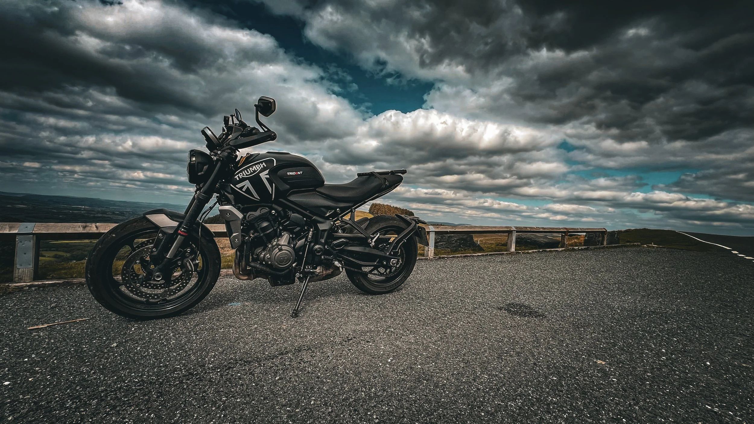 Black Triumph motorcycle parked on the side of a mountain road with a cloudy sky overhead.