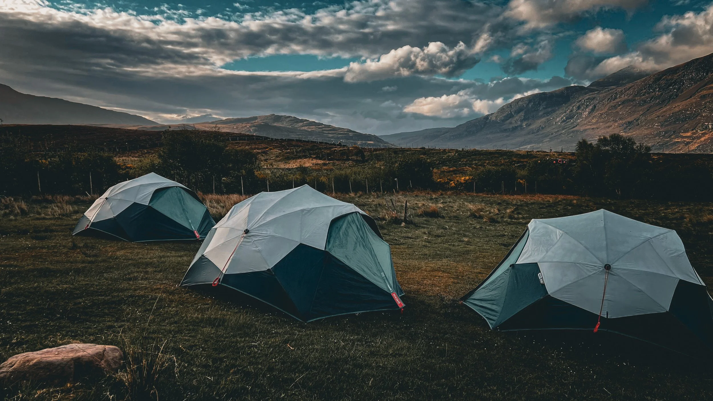 Four camping tents set up on grassy land with mountain ranges and cloudy sky in the background