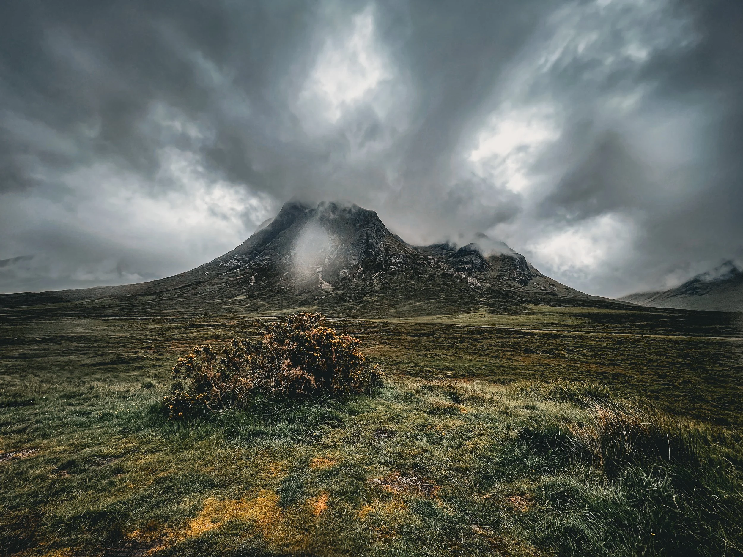 A rugged mountain peak with storm clouds overhead and a green grassy field in the foreground.