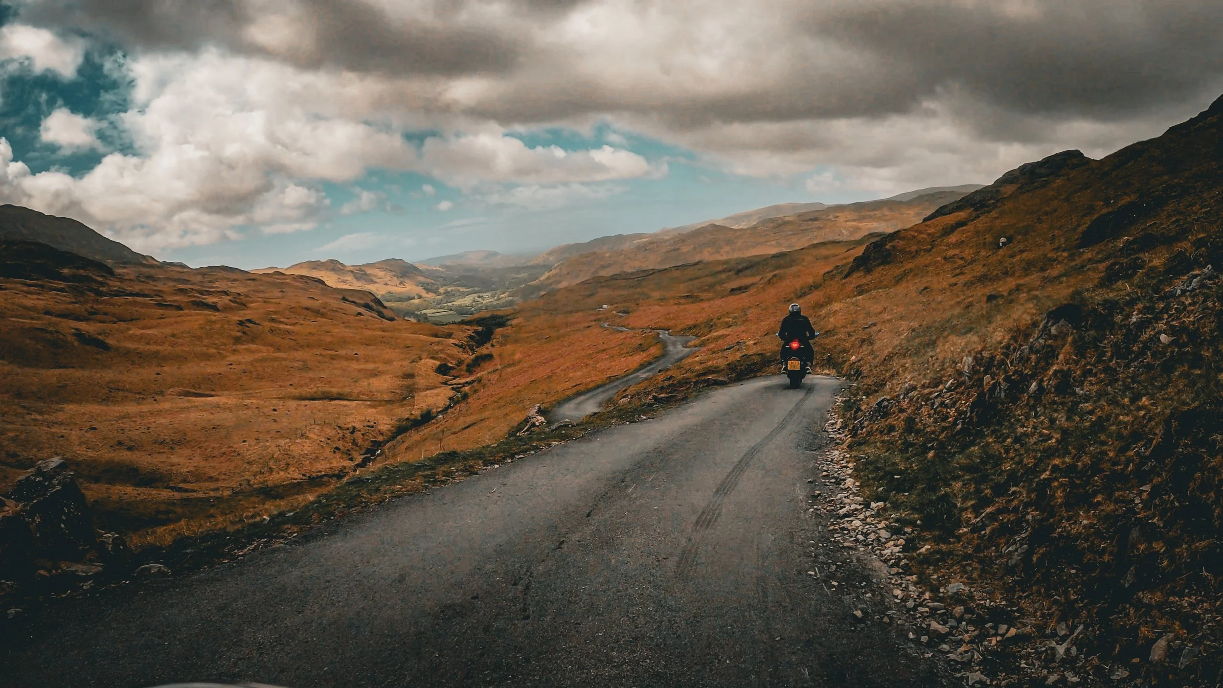 A person riding a motorcycle down a narrow winding mountain road surrounded by brown hills under a cloudy sky.