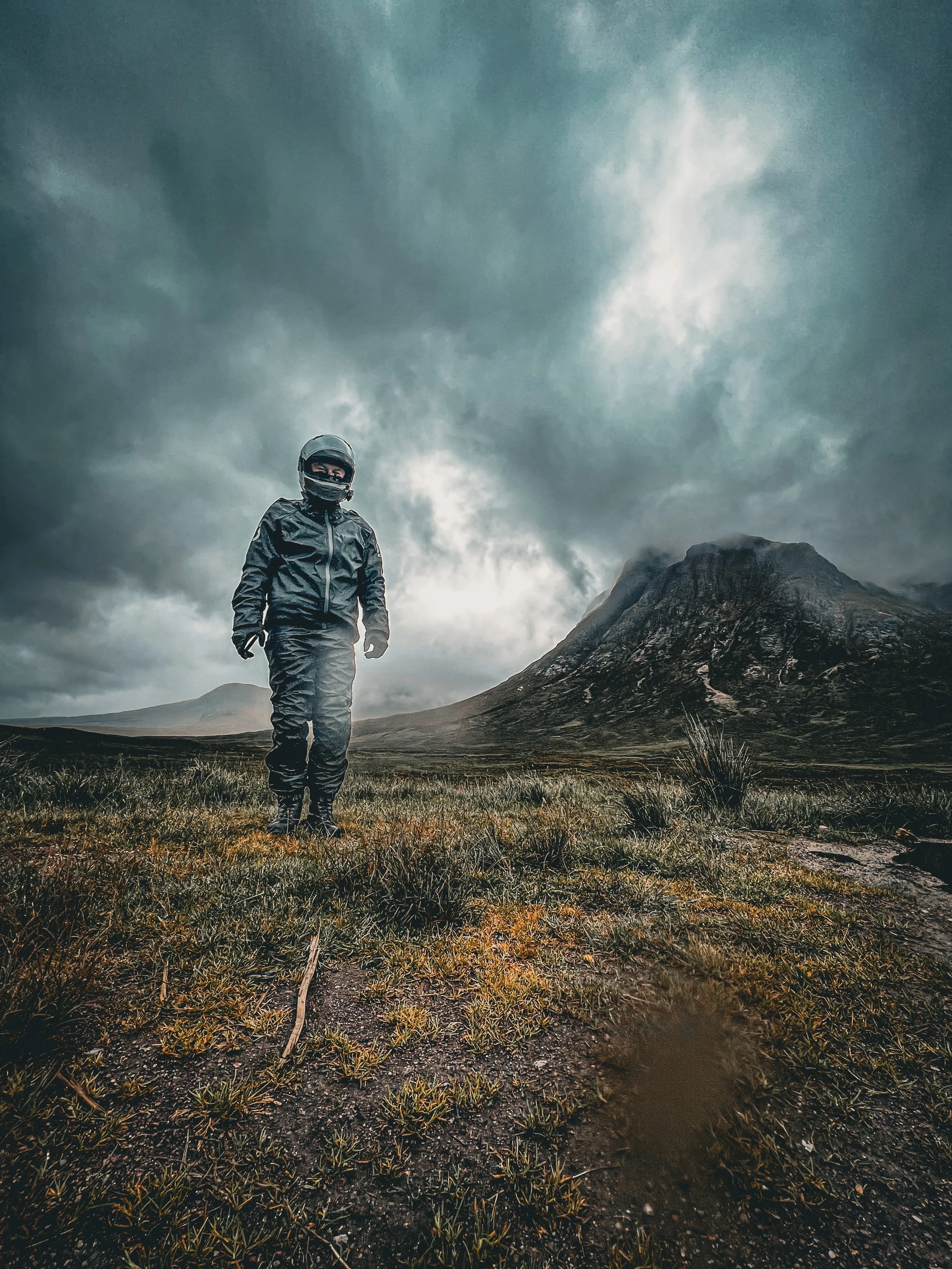 A person wearing motorcycle gear, including a helmet and jacket, standing on a grassy terrain in front of a mountain with dark, cloudy skies overhead.