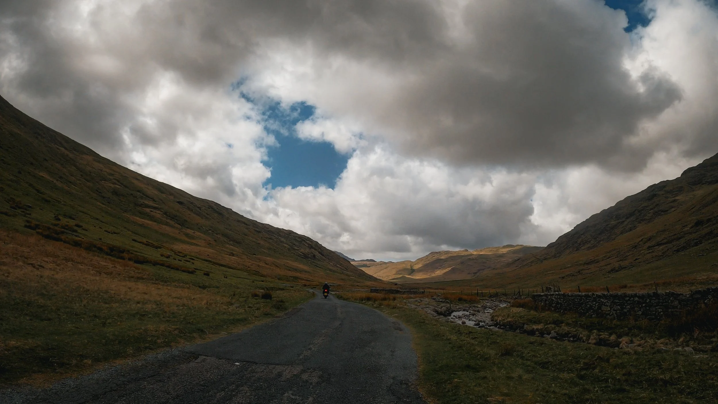 Scenic view of a valley with a winding road, surrounded by green rolling hills and mountains, under a cloudy sky with patches of blue visible.
