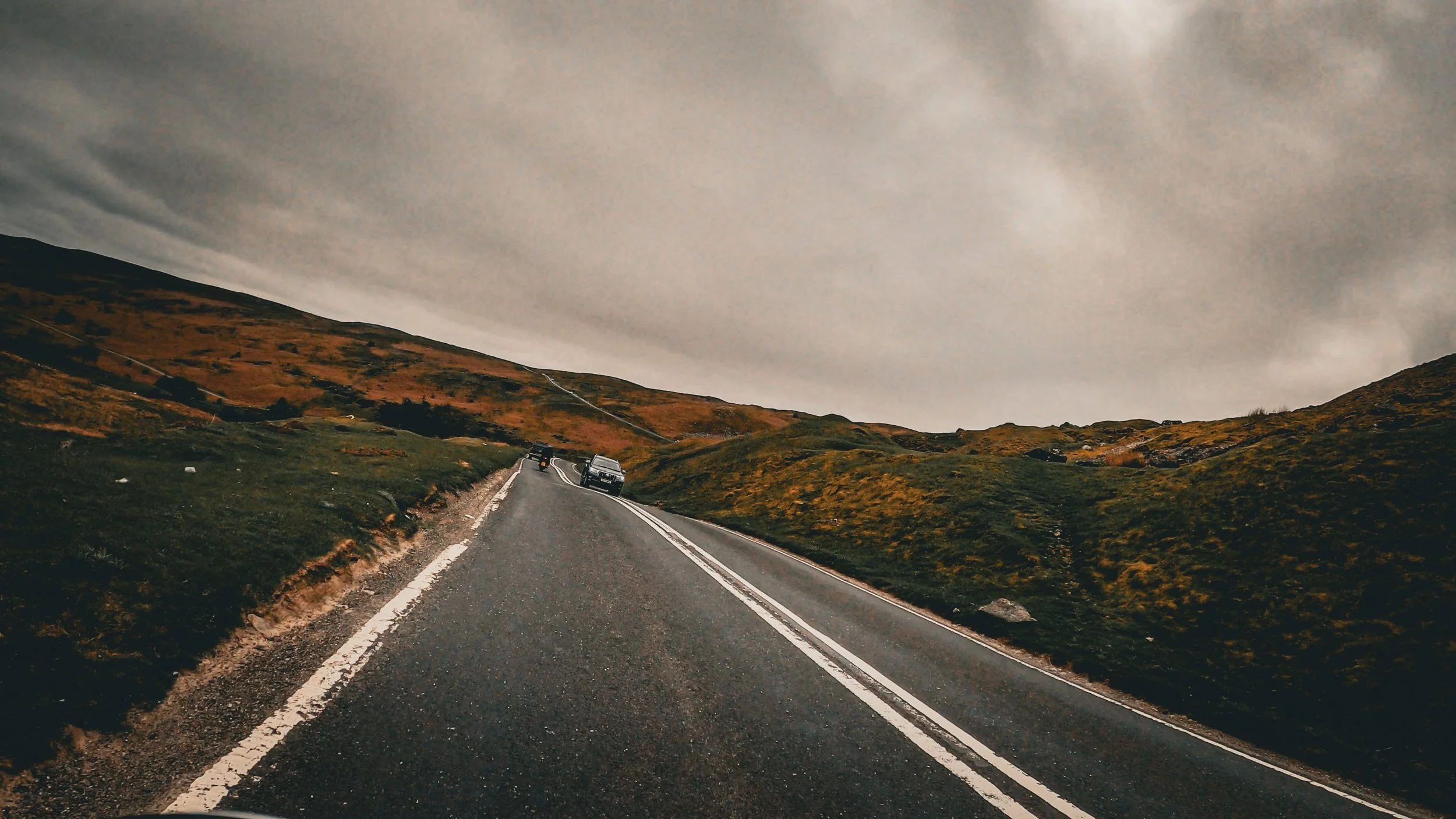 A winding mountain road with a few cars, surrounded by green and brown hills, under a cloudy sky.