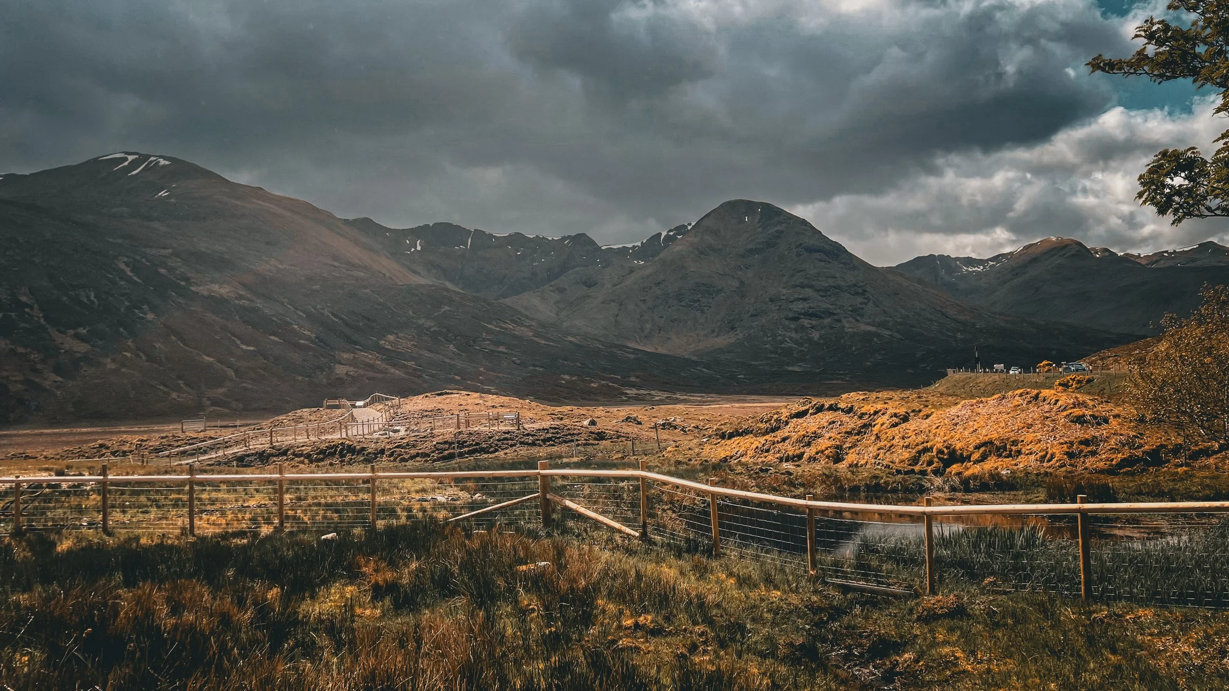 A mountainous landscape with dark, cloudy skies overhead, a fenced pathway in the foreground, and a small group of buildings on the right side of the image.