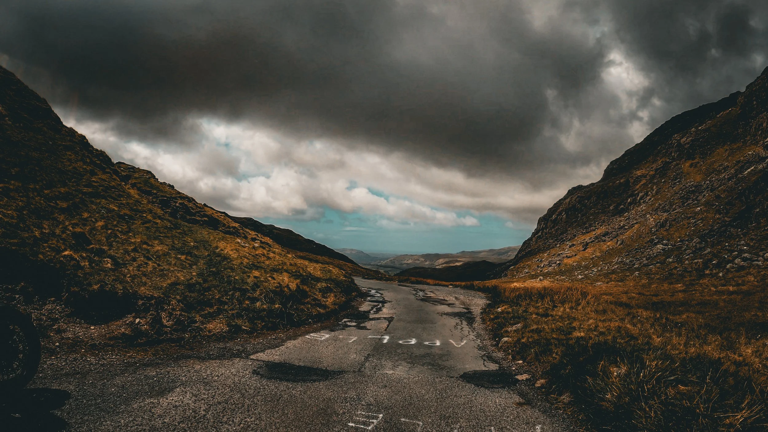 A winding mountain road with cracked pavement, flanked by steep hills covered in grass and rocks, under a dark, cloudy sky.