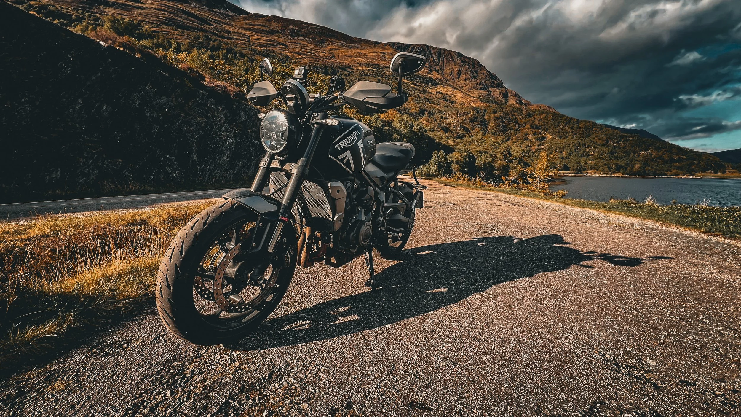 A black Triumph motorcycle parked on a gravel road near a body of water, with mountains covered in trees and a cloudy sky in the background.
